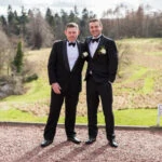 groom and best man wearing black tuxedos and bow ties standing outside the castle