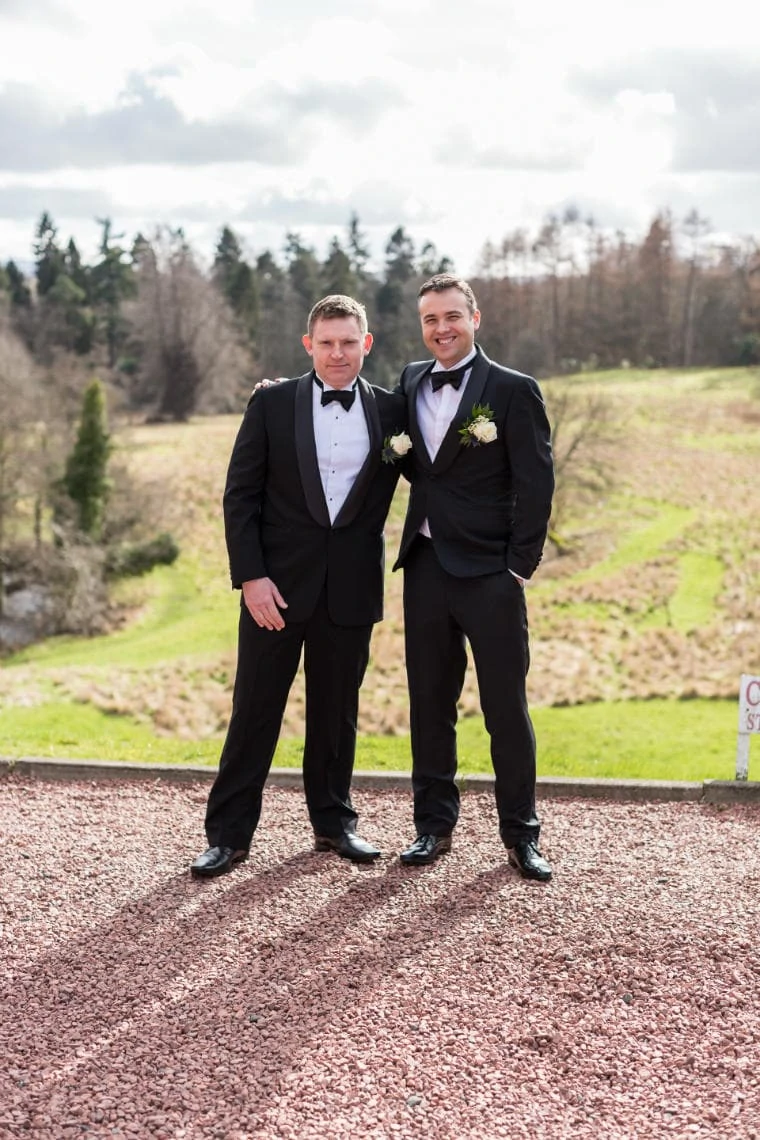 groom and best man wearing black tuxedos and bow ties standing outside the castle