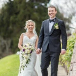 newlyweds smiling at the camera as they walk down the path towards the arched bridge