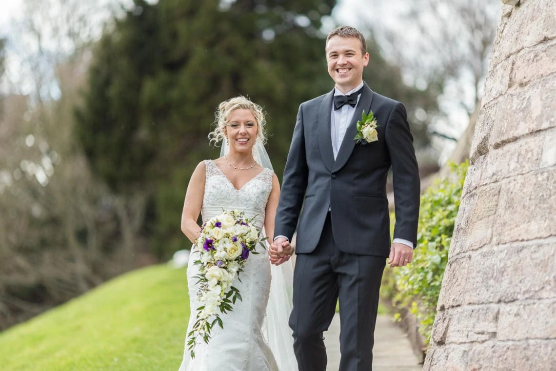 newlyweds smiling at the camera as they walk down the path towards the arched bridge