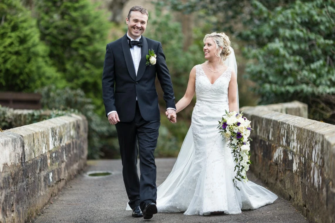 newlyweds smiling and holding hands as they walking across the bridge