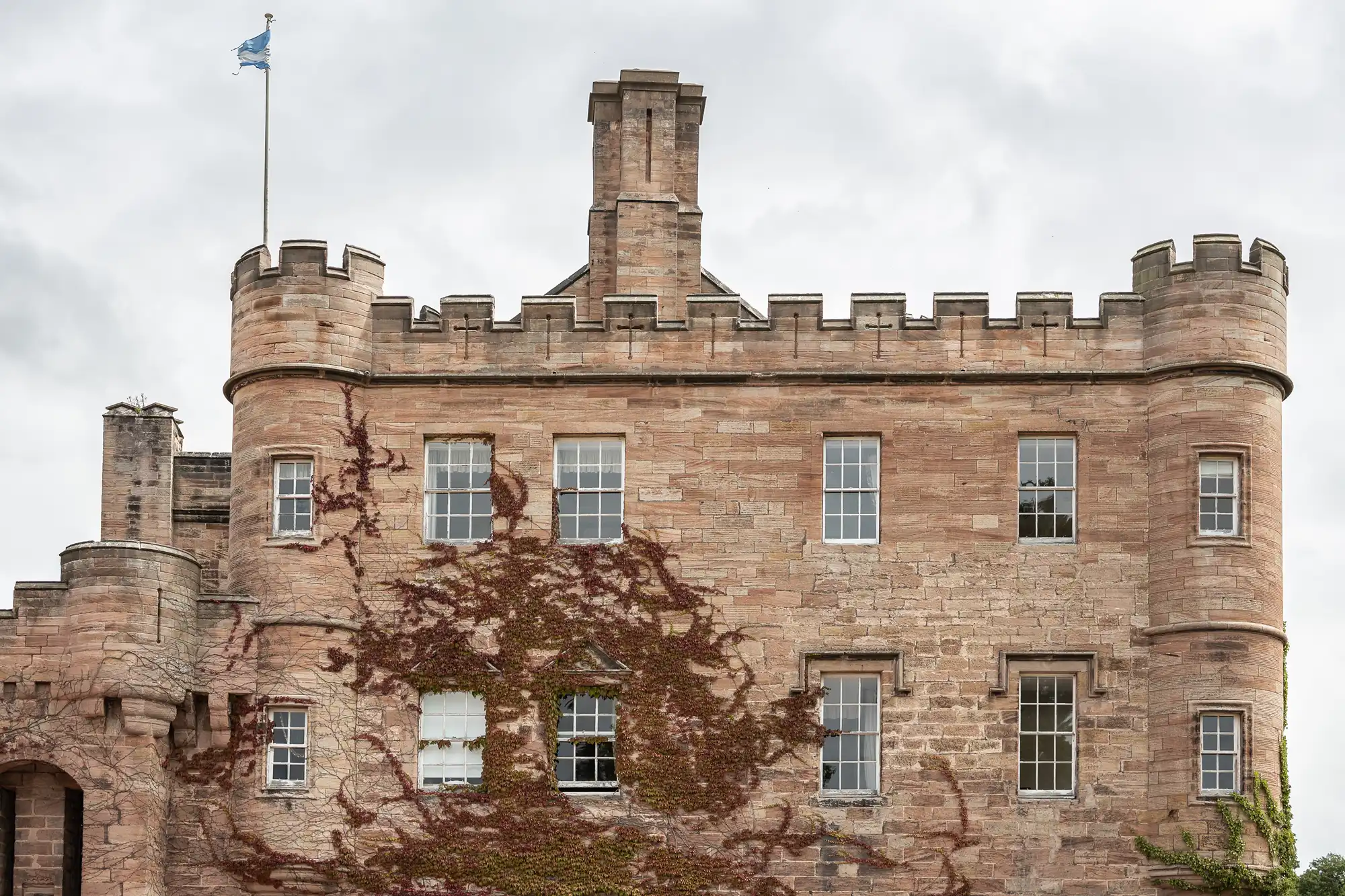 A stone castle with ivy climbing the walls is shown. It has round turrets with crenellations, several windows, and a flag flying on top. The sky is overcast.