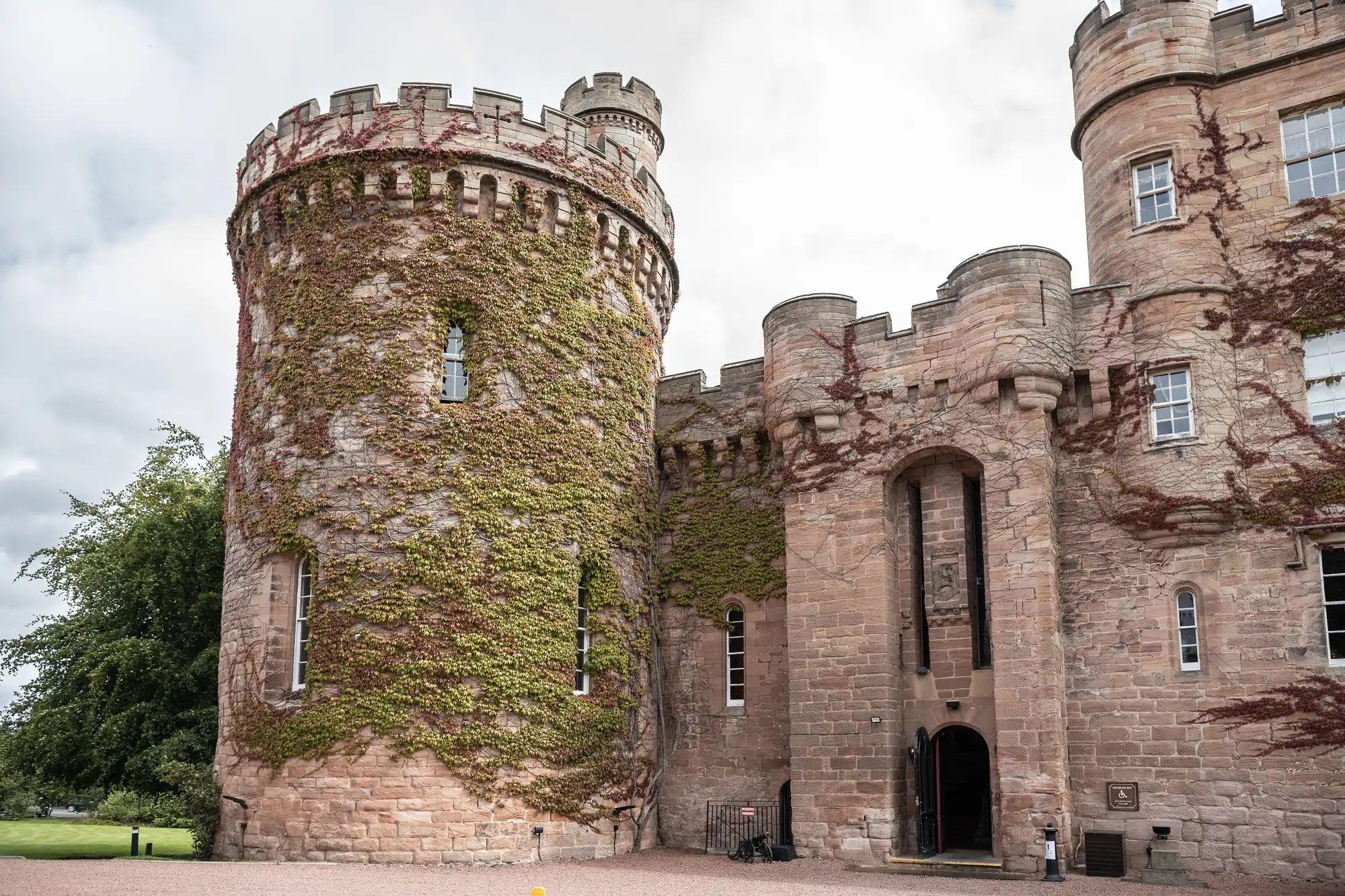 A large stone castle with a cylindrical turret covered in ivy, featuring narrow windows and an entryway below a tall arch, with a gravel area in front.