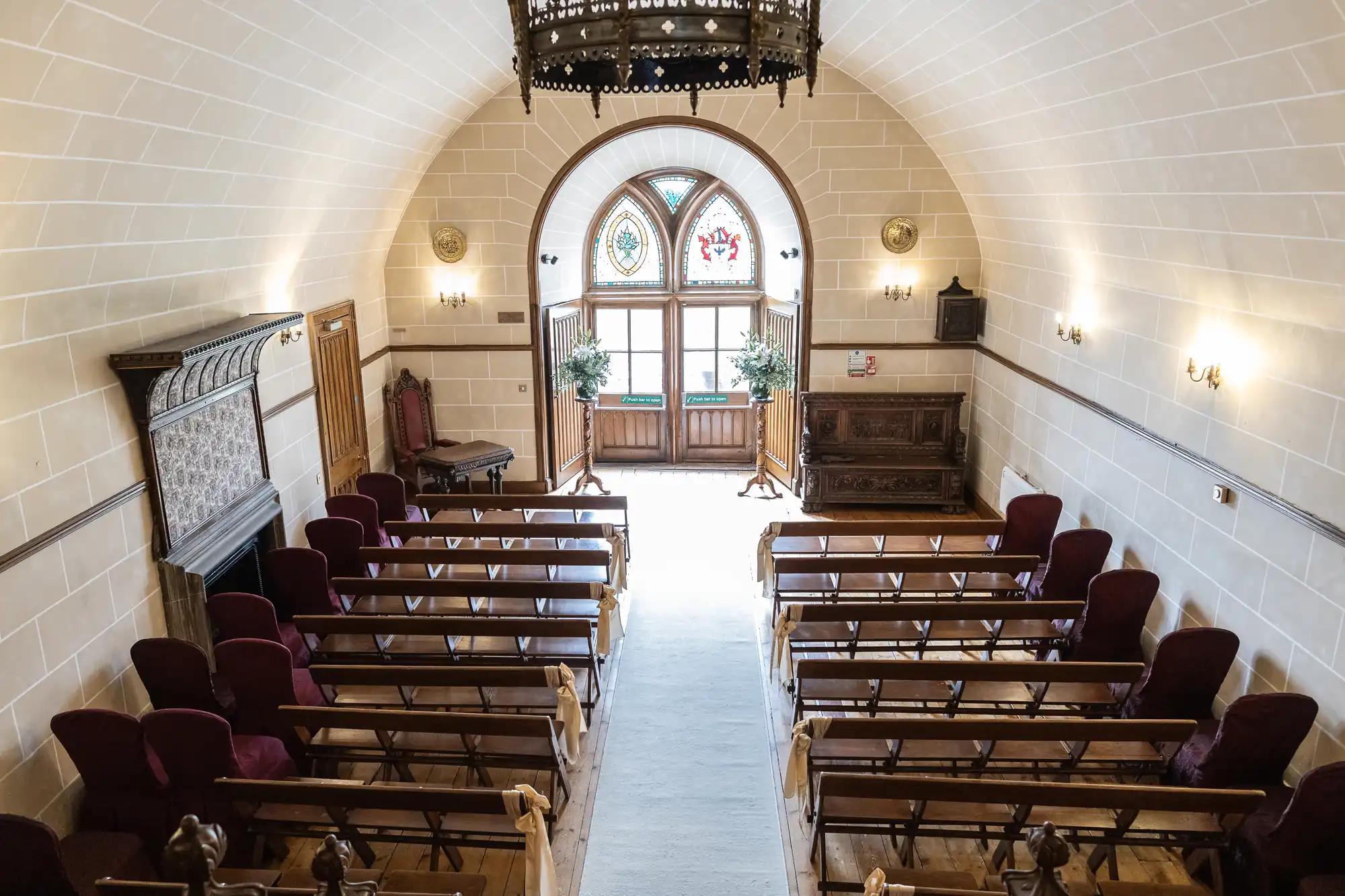 A small chapel with rows of wooden pews, arched windows, a chandelier, and light-colored walls. The room is lit with natural light and wall sconces.