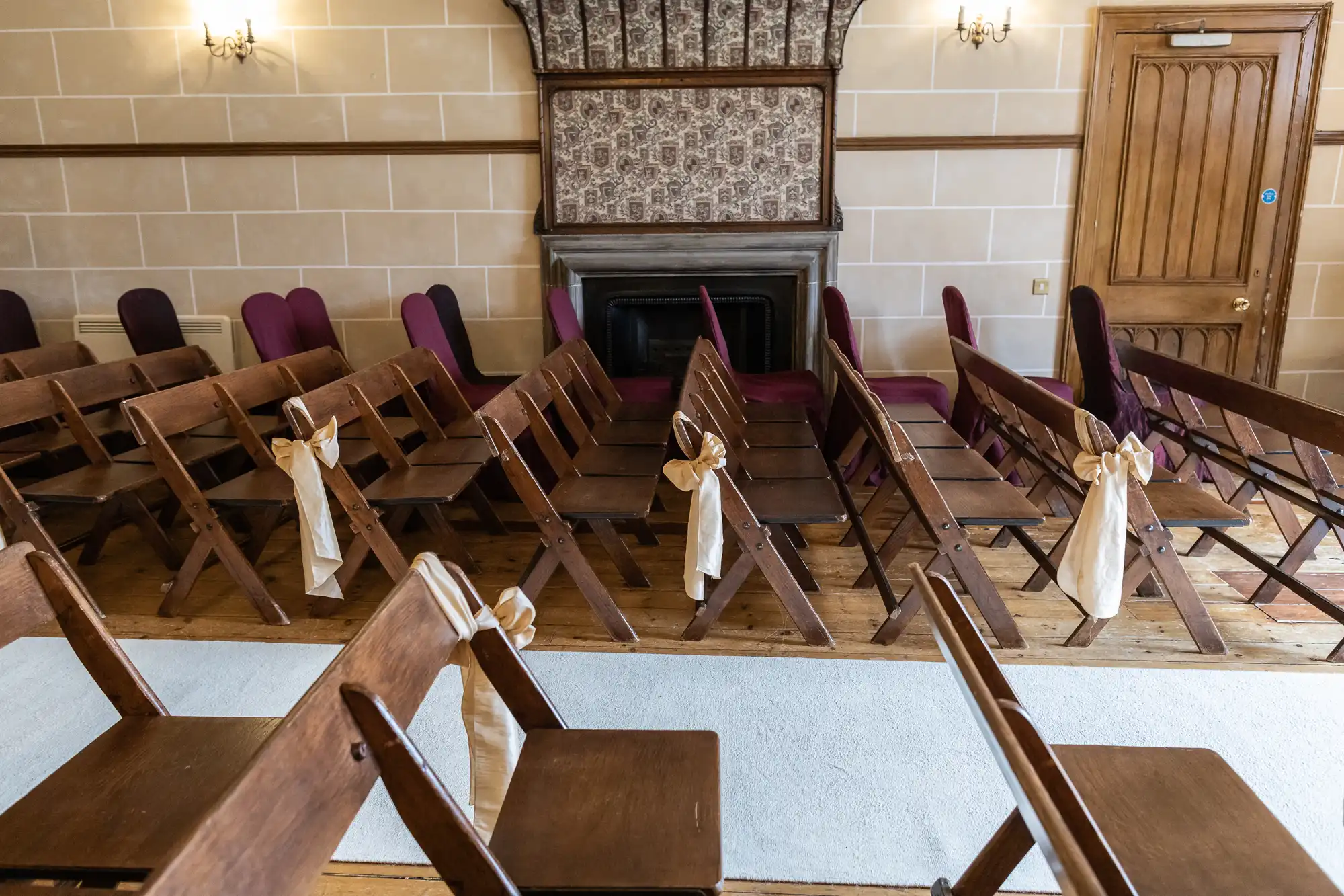 Rows of wooden chairs decorated with beige ribbons are set up facing a fireplace in a room with beige walls and a wooden door.