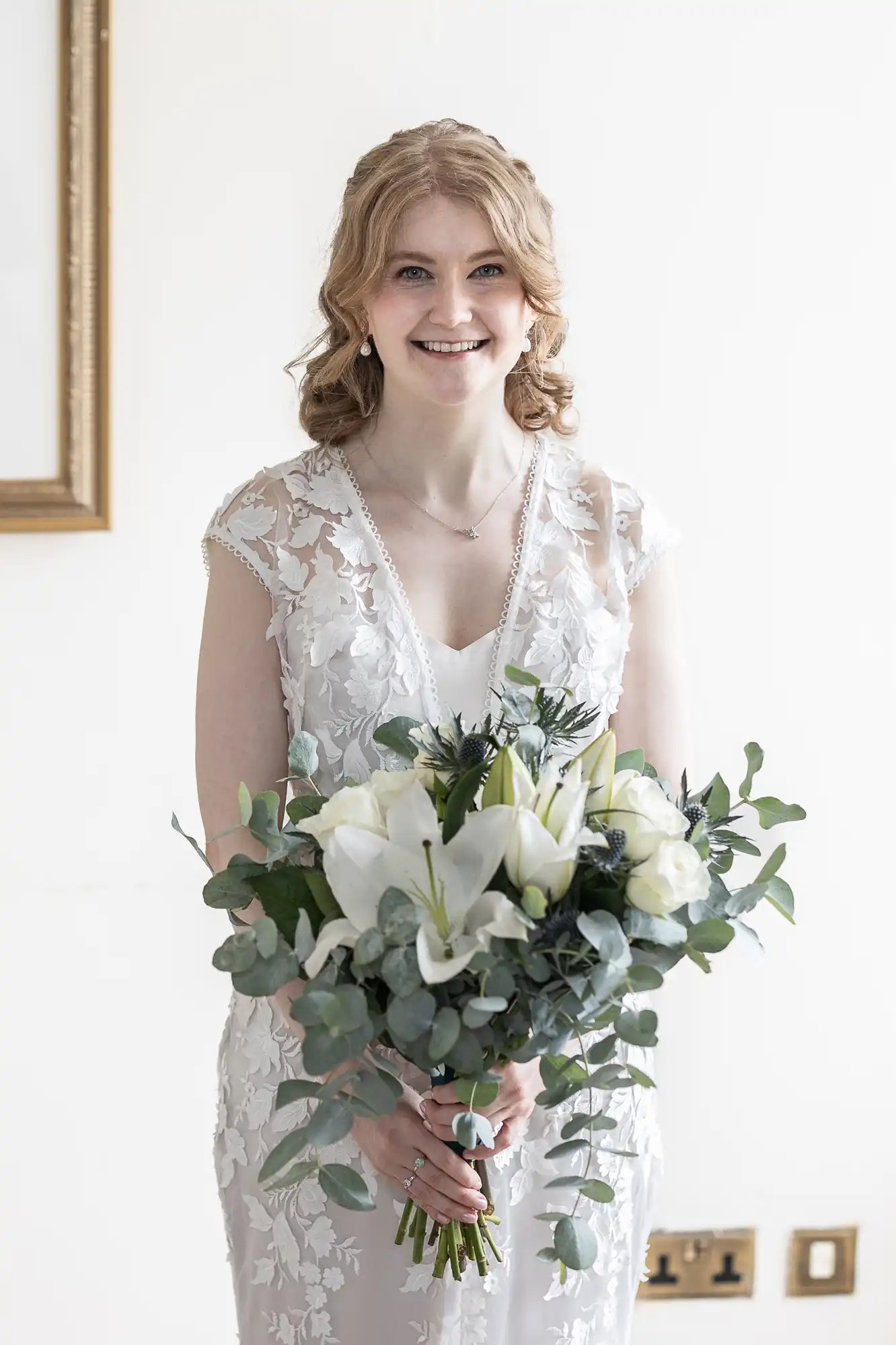 A woman in a white lace wedding dress holds a bouquet of white and green flowers, standing against a light background.