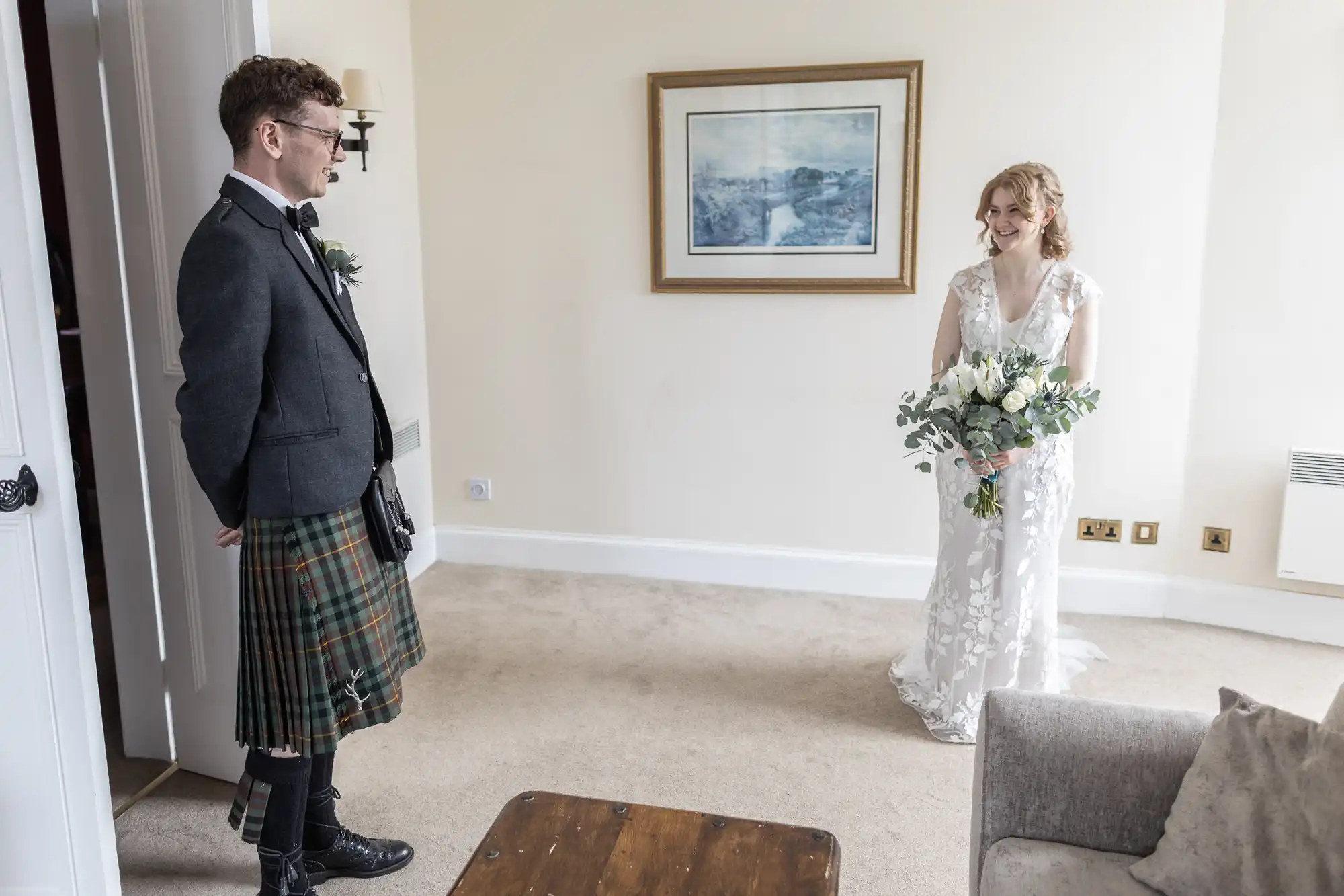 A man in a kilt and a woman in a wedding dress stand in a living room, looking at each other. The woman holds a bouquet, and a painting hangs on the wall behind them.