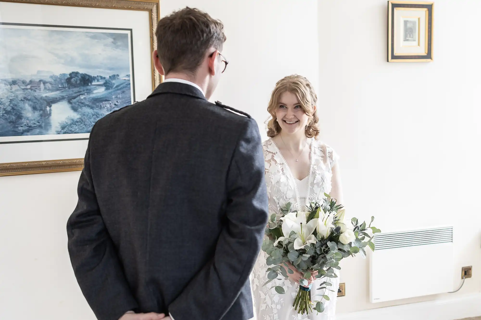 A bride holding a bouquet of white flowers smiles at a man in a suit. They are indoors with a framed painting in the background.