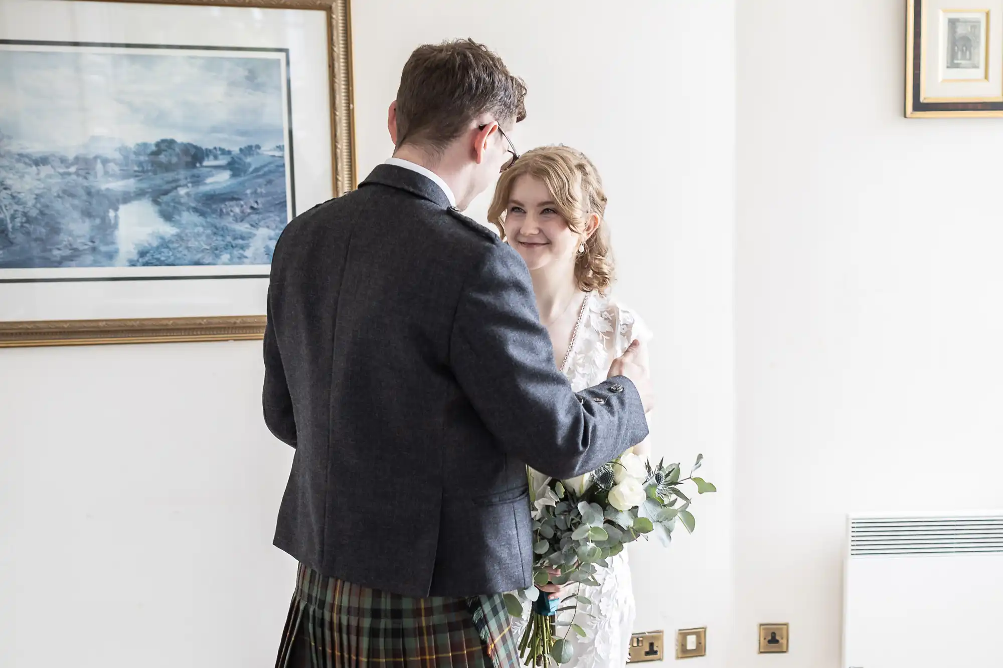 A couple faces each other; the man wearing a kilt and jacket holds the woman's arm as she smiles. She holds a bouquet of greenery and flowers. They stand indoors, with framed pictures on the wall behind them.