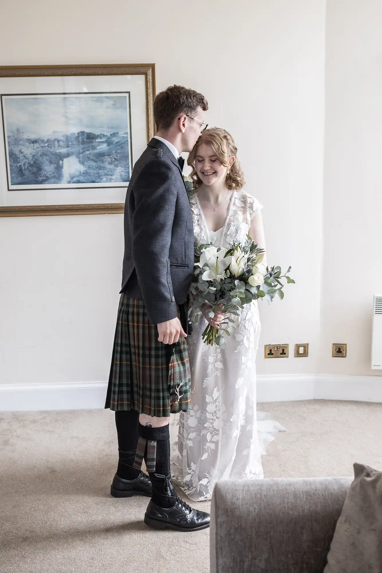 A couple stands together indoors. The man wears a kilt and a jacket, and the woman wears a white gown and holds a bouquet. They are smiling and facing each other. A framed picture hangs on the wall behind them.