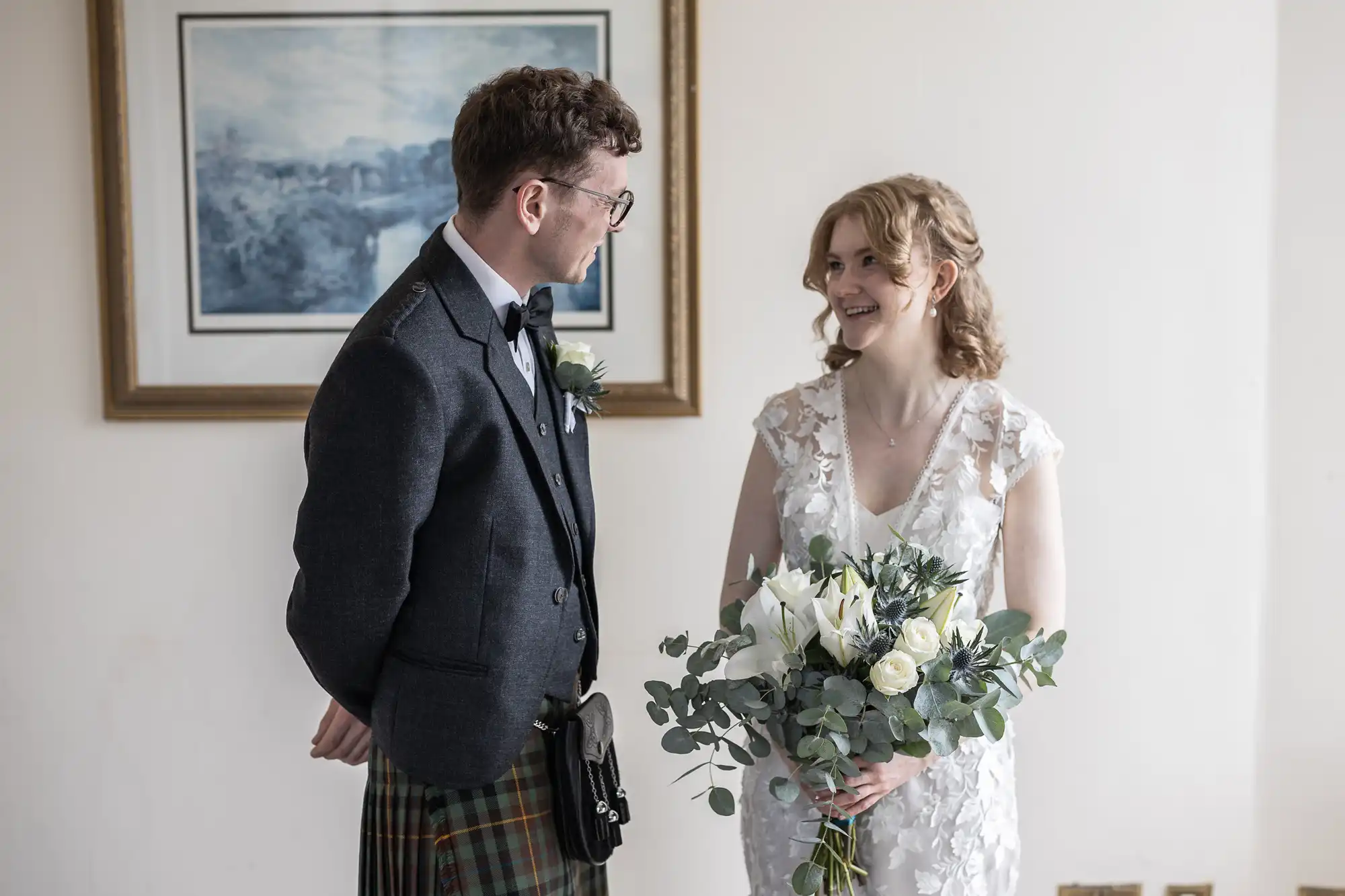 A couple stands together indoors, the man in a dark suit with a kilt and the woman in a white dress holding a bouquet, both smiling at each other. A framed picture hangs on the wall behind them.