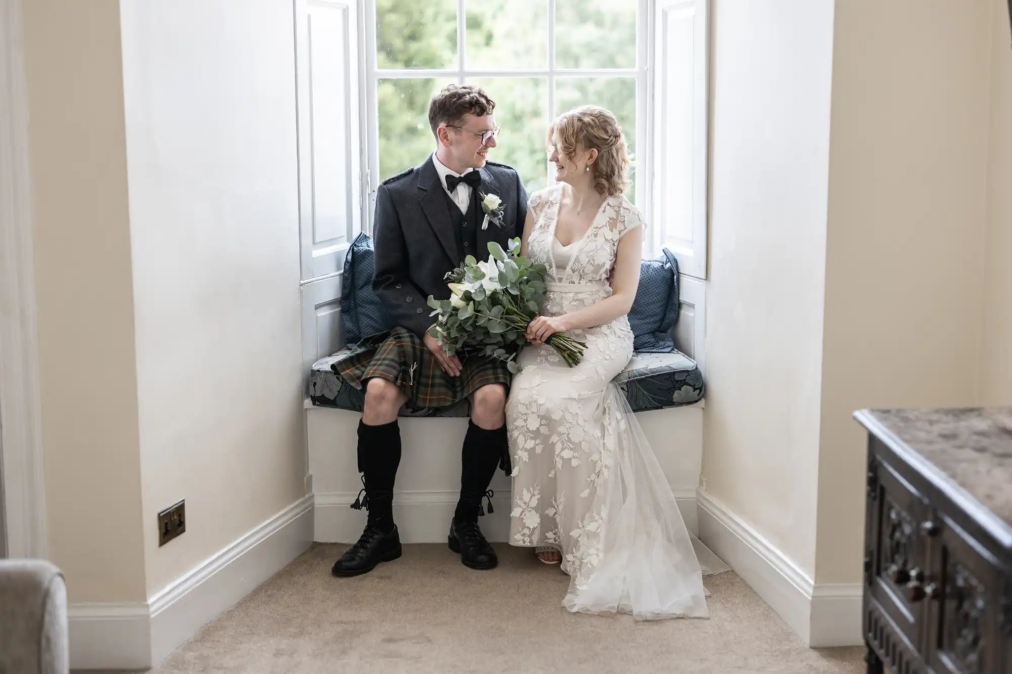 A couple dressed in wedding attire sits on a window seat. The groom is in a kilt, and the bride holds a bouquet of greenery. They face each other, smiling softly.