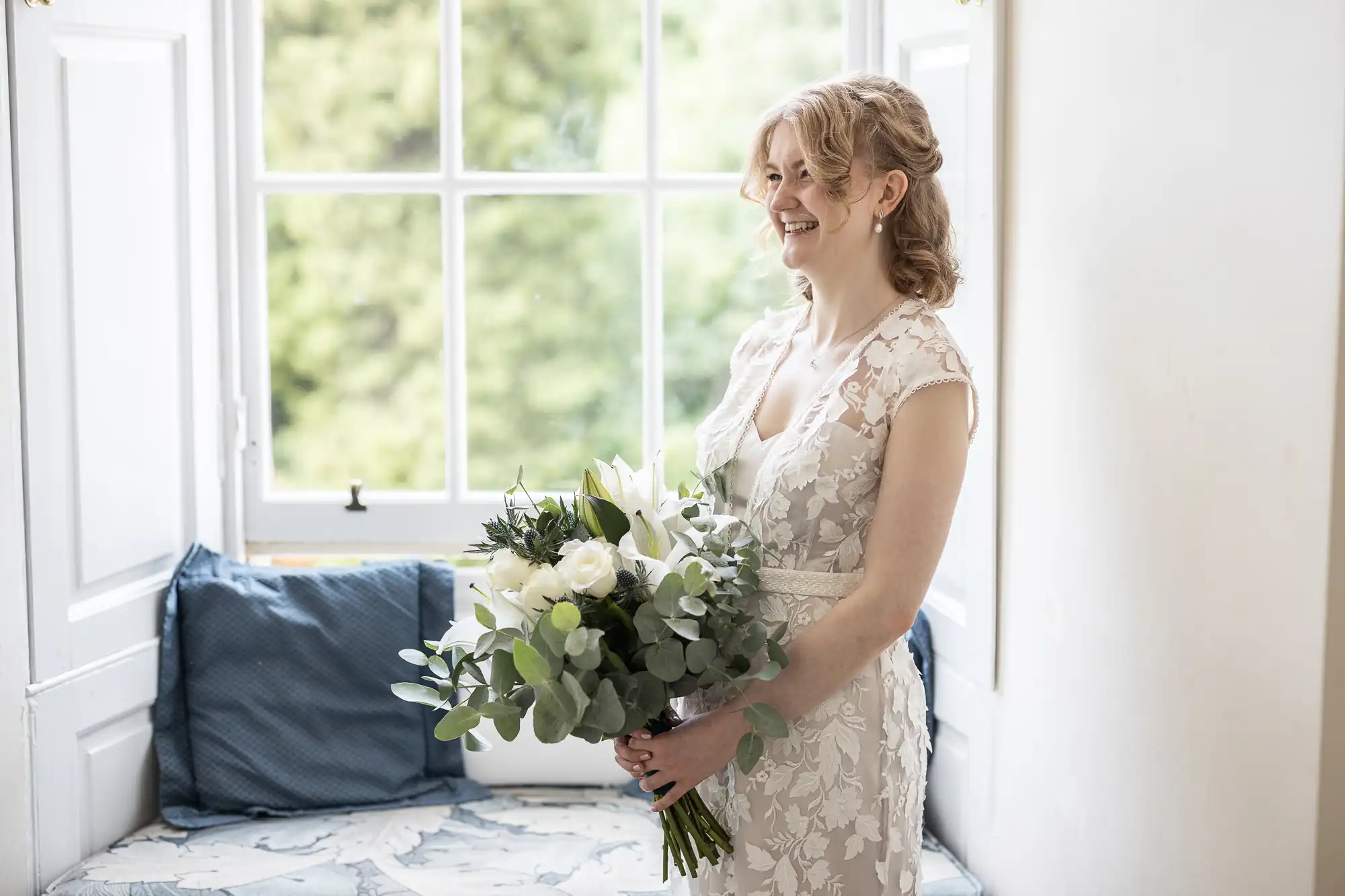 A woman in a lace dress stands by a window holding a bouquet of flowers, smiling. She has blonde hair and is in a bright room with a window seat and blue cushions.