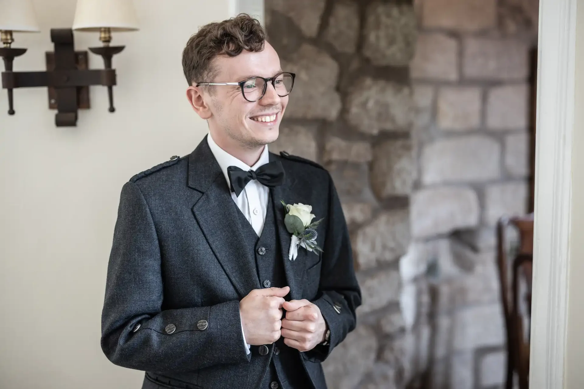 A person wearing glasses and a dark suit with a bow tie and boutonniere stands smiling near a stone wall and sconce light.
