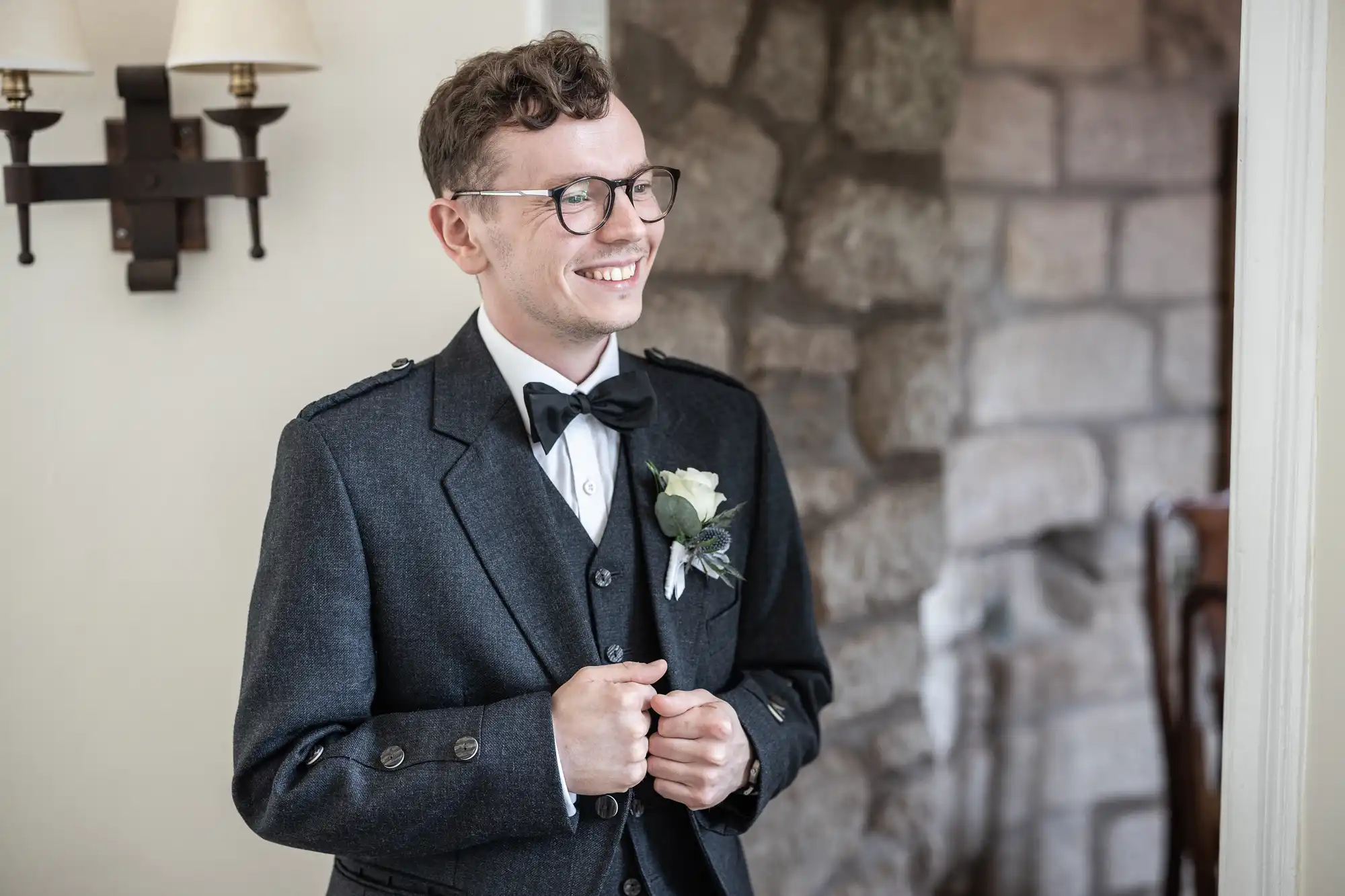 A person wearing glasses and a dark suit with a bow tie and boutonniere stands smiling near a stone wall and sconce light.