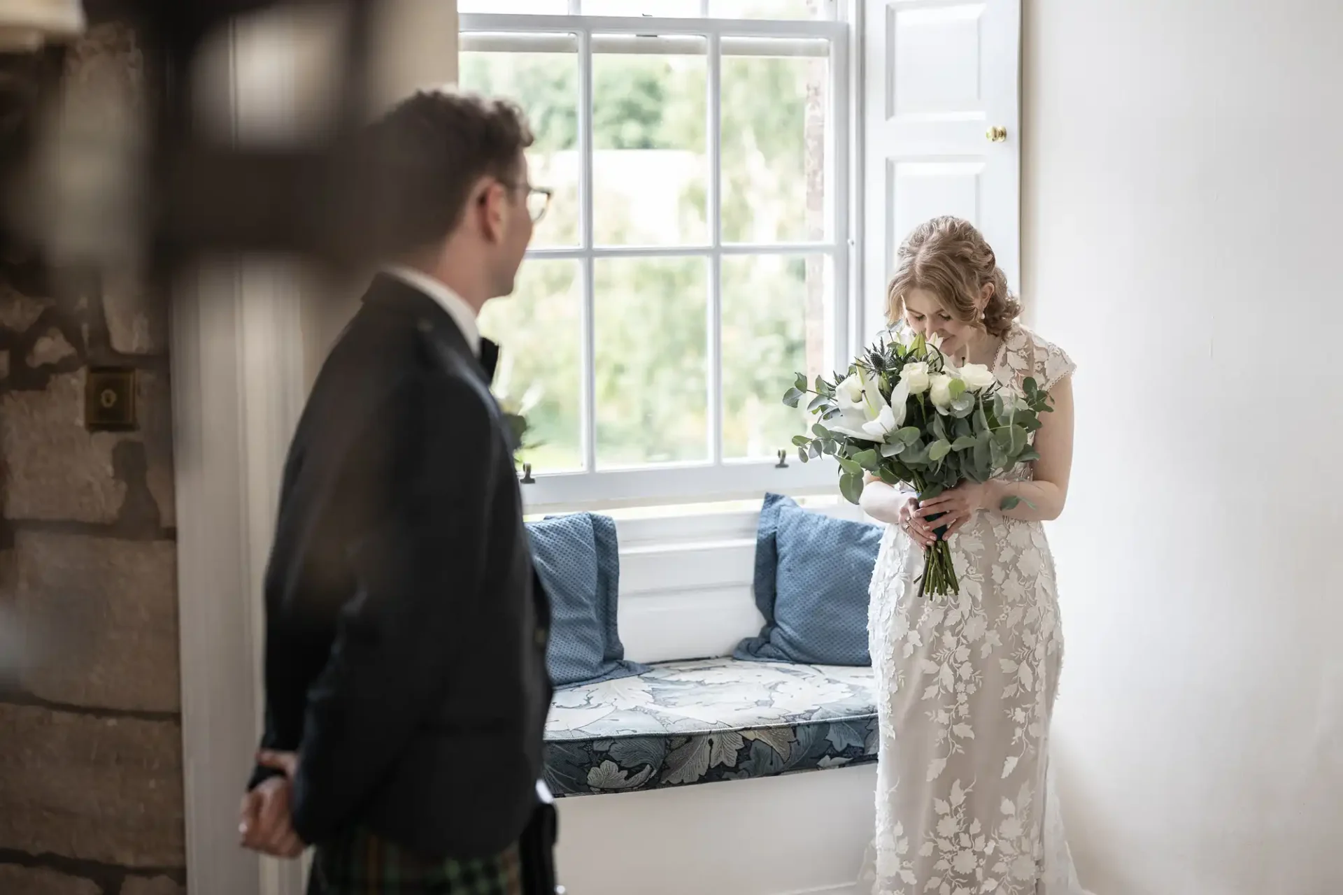 A woman in a white dress holds a bouquet of flowers by a window, while a man in formal attire stands nearby.