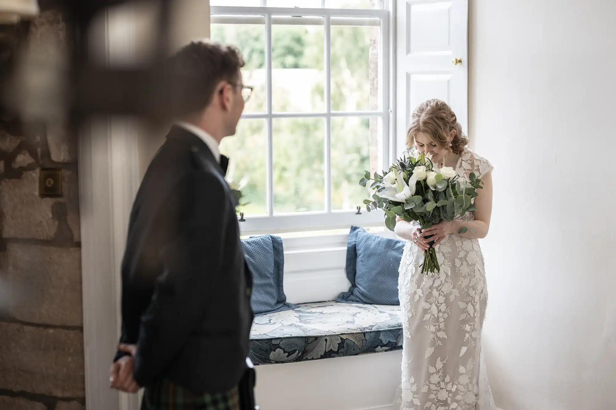 A woman in a white dress holds a bouquet of flowers by a window, while a man in formal attire stands nearby.