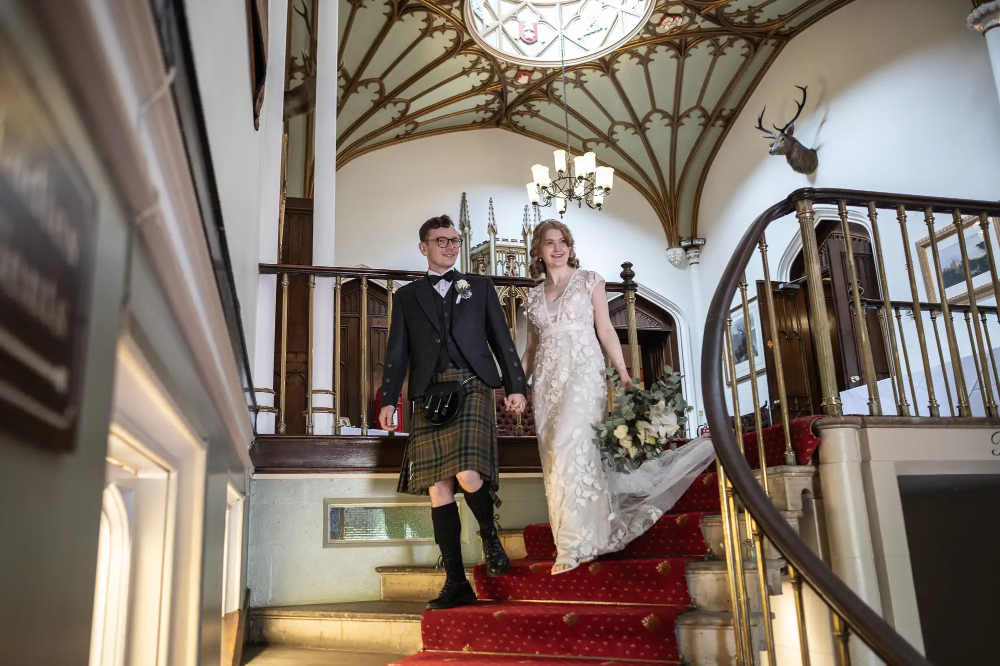 A couple descends a red-carpeted staircase in a historic venue. The groom wears a kilt and the bride is in a white gown, holding a bouquet. The interior features ornate ceilings and a chandelier.