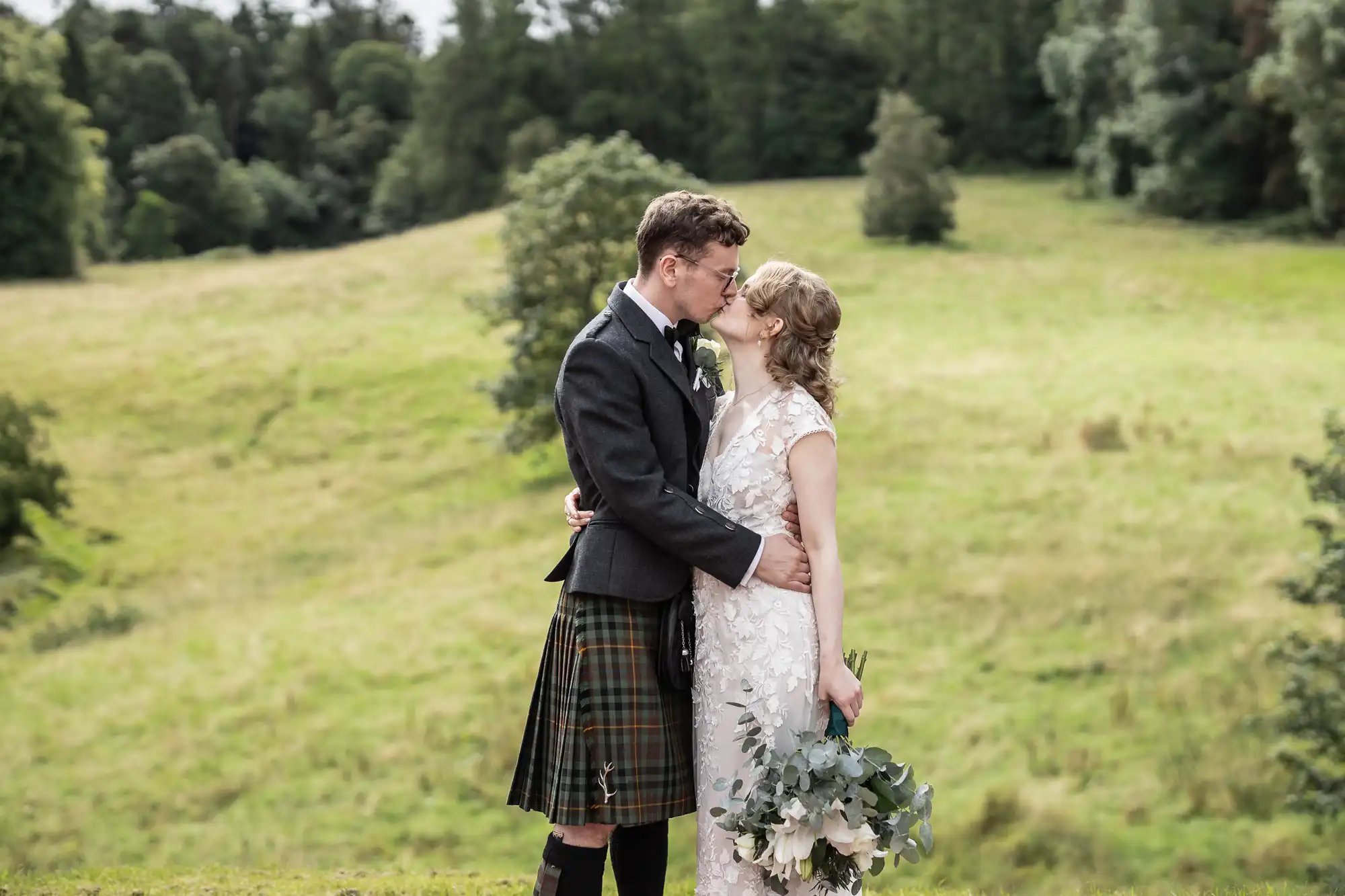 A couple is kissing outdoors on a grassy hill. The man is wearing a kilt and jacket, while the woman is dressed in a white gown holding a bouquet. Trees are visible in the background.