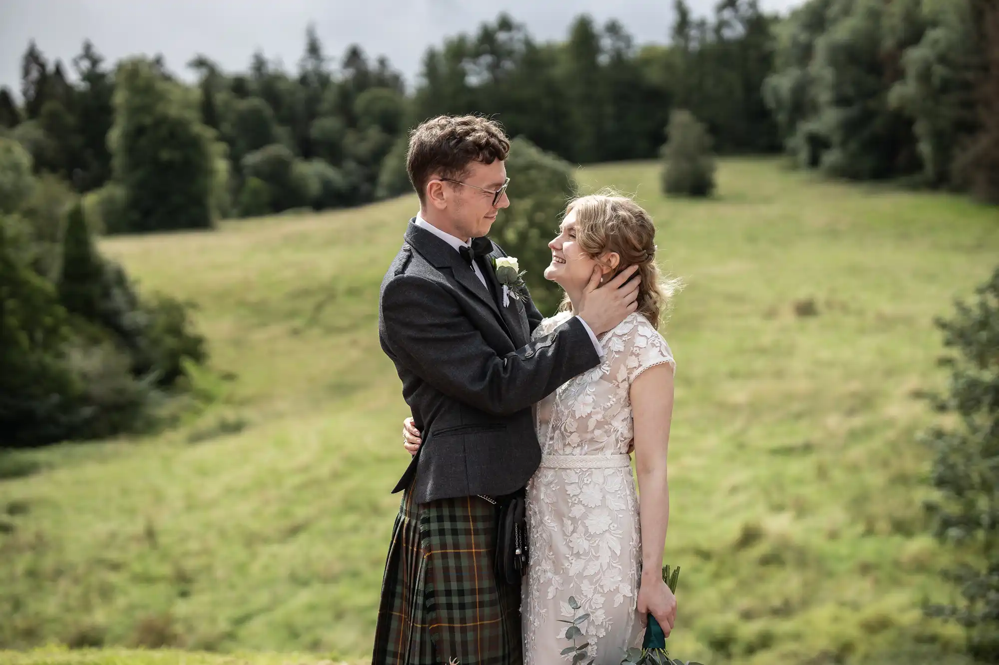 A couple dressed in formal attire stands closely together in a grassy, hilly outdoor setting, with the man in a kilt and the woman in a white dress.