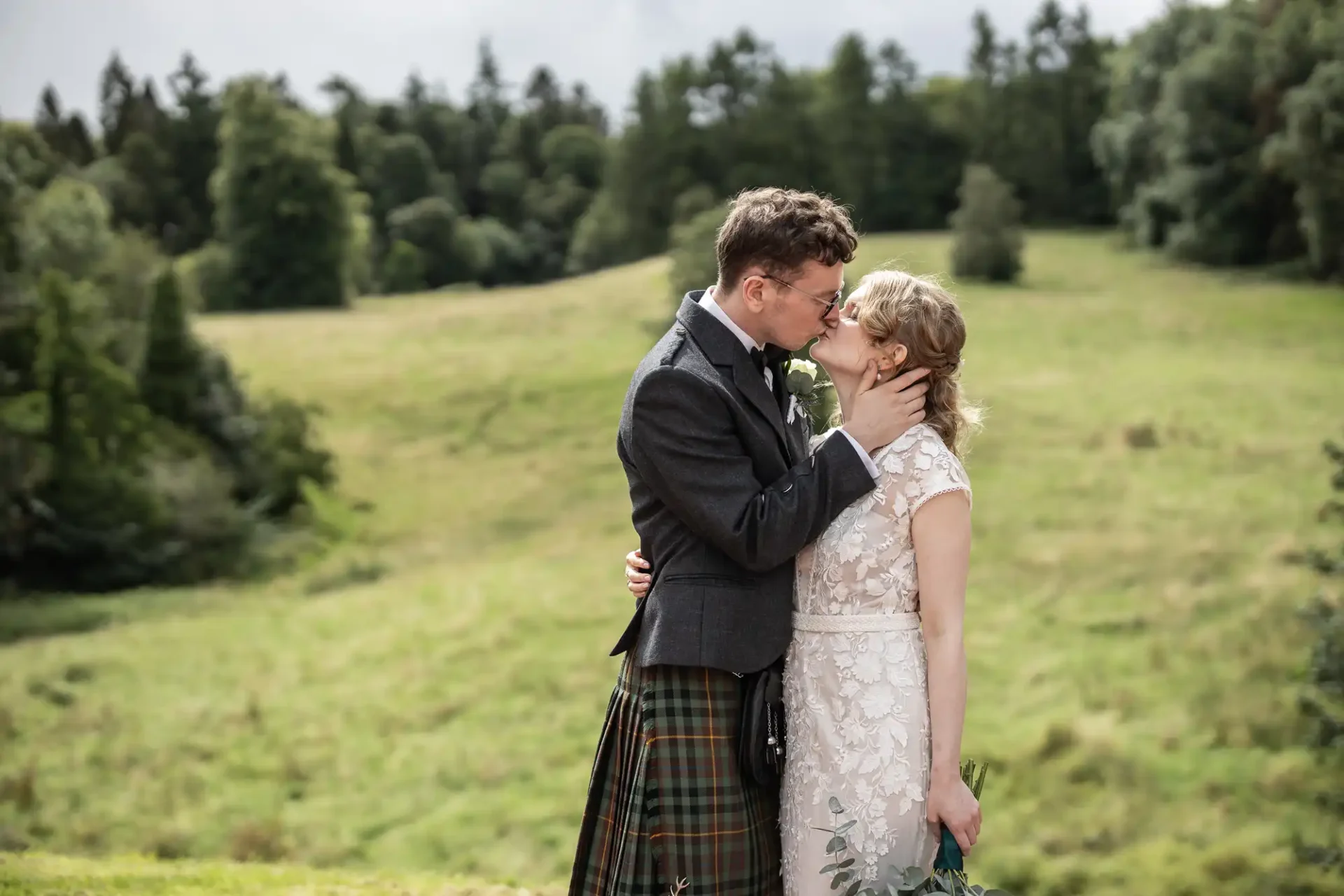 A groom in a kilt kisses the bride on the forehead as they embrace in a grassy, wooded outdoor setting.