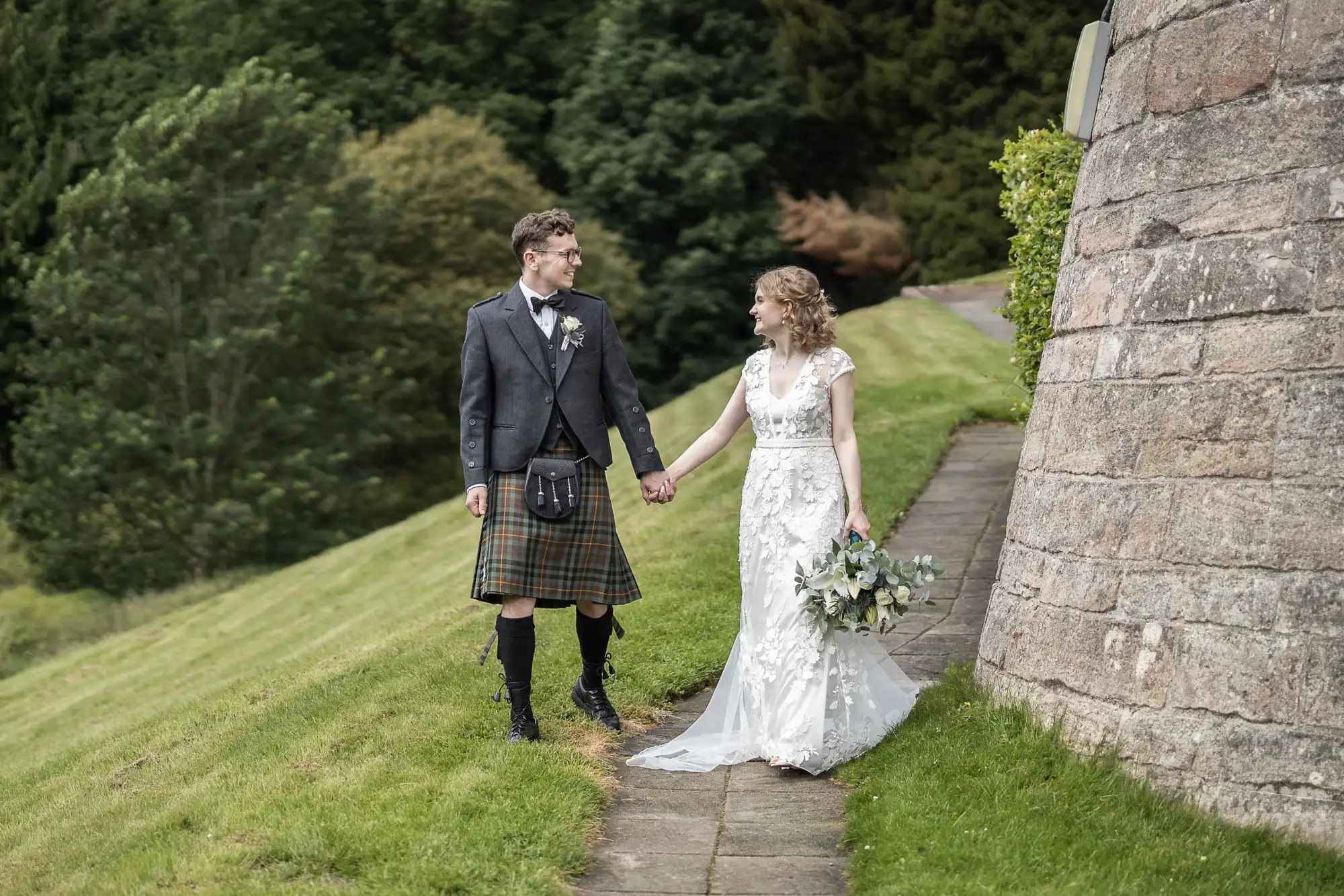 A couple, with the man in a kilt and the woman in a white wedding dress, walk hand-in-hand on a stone path beside a stone wall and lush greenery.