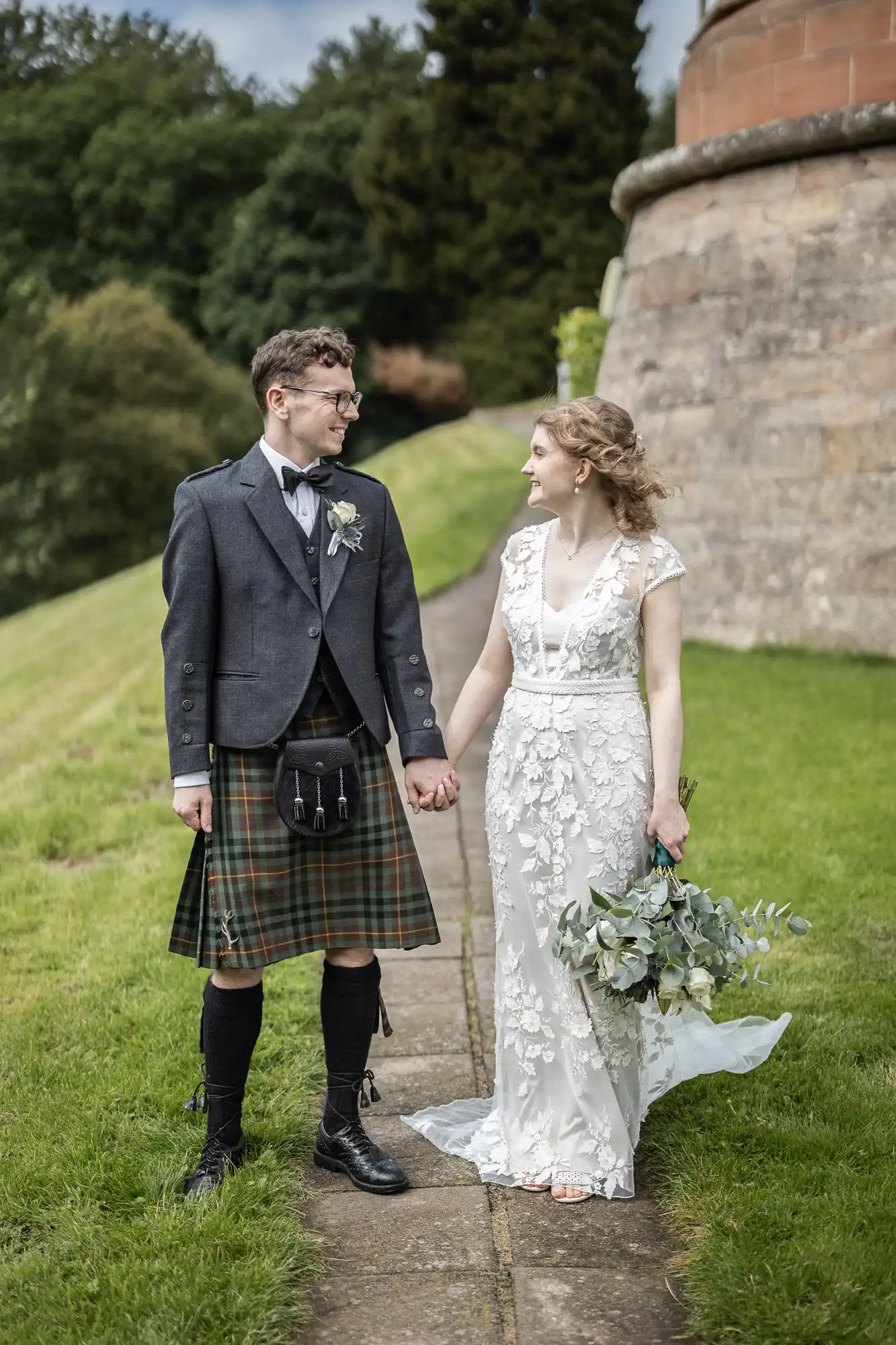 A couple stands outdoors holding hands. The man is dressed in a kilt and jacket, while the woman wears a white lace gown and holds a bouquet. They are on a pathway next to a stone building with greenery in the background.