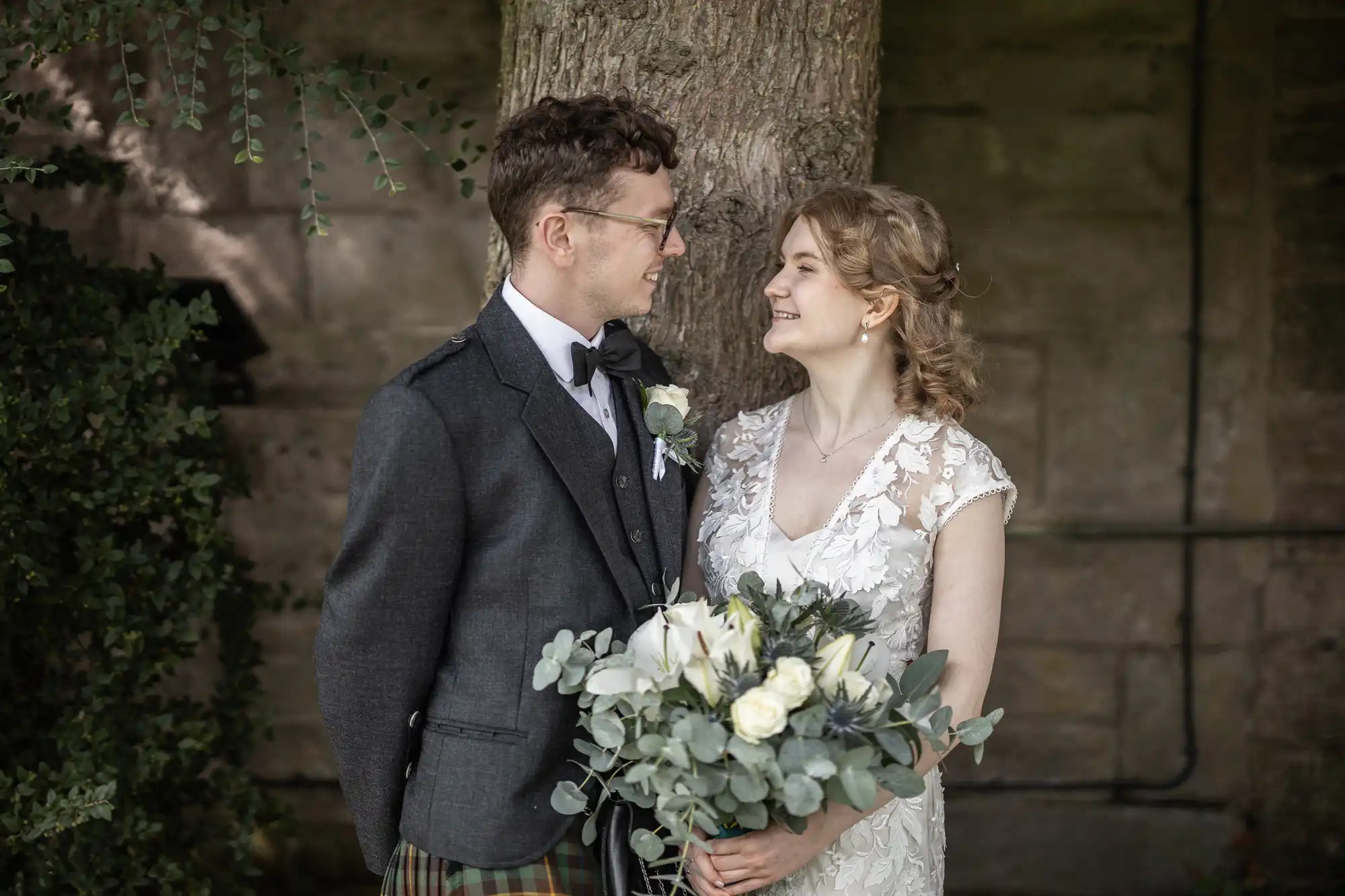 A bride and groom, dressed in wedding attire, stand close together near a tree. The bride holds a bouquet of white flowers and eucalyptus. They are looking at each other, smiling.