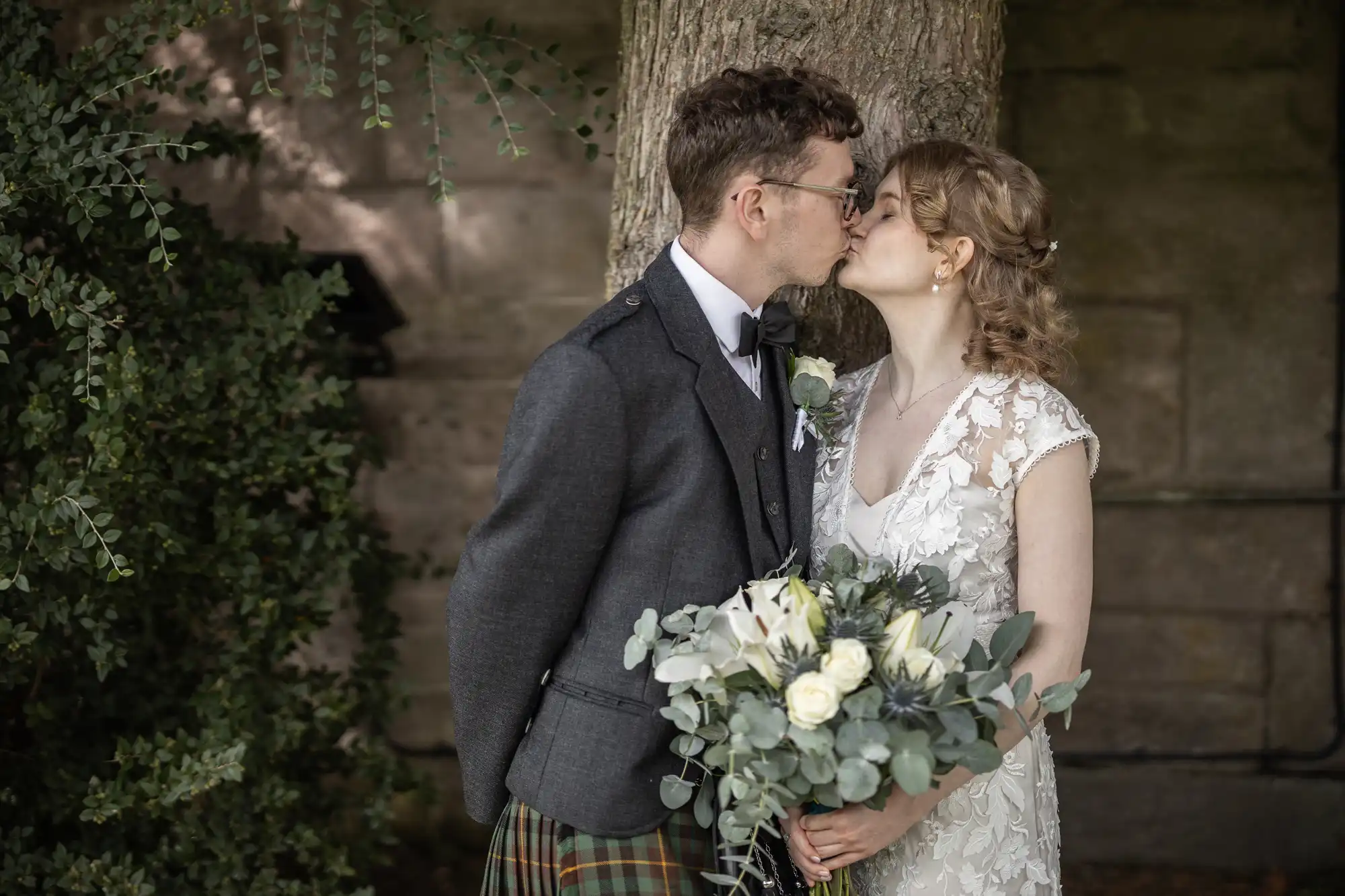 A couple shares a kiss while dressed in wedding attire; the groom wears a gray suit jacket and tartan pants, and the bride wears a white lace dress and holds a bouquet of white flowers and greenery.