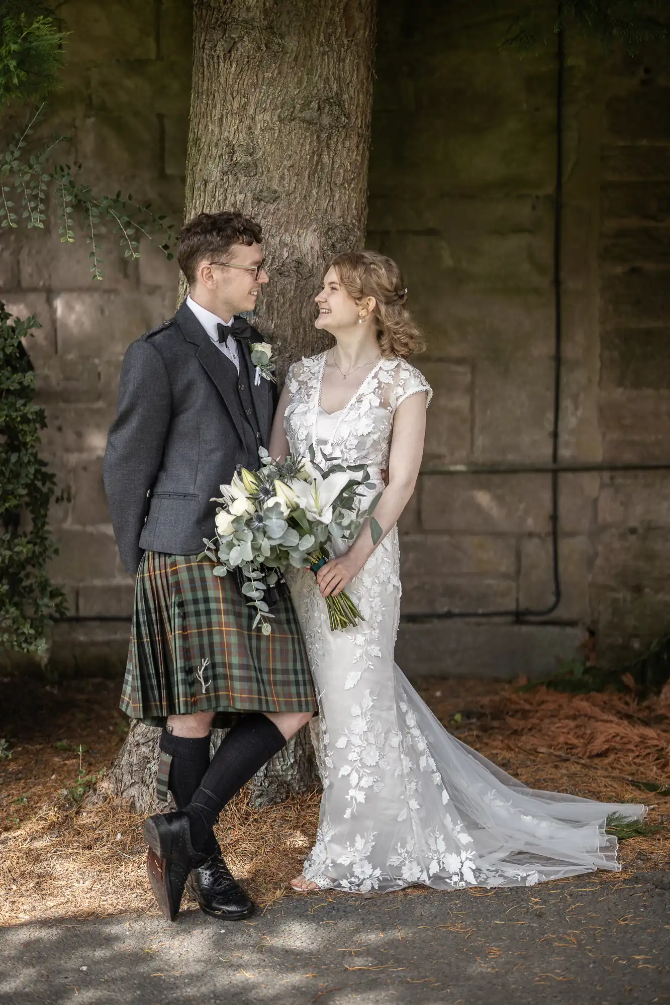 A couple stands next to a tree, the man in a kilt and jacket, the woman in a white floral dress holding a bouquet, both looking at each other and smiling.