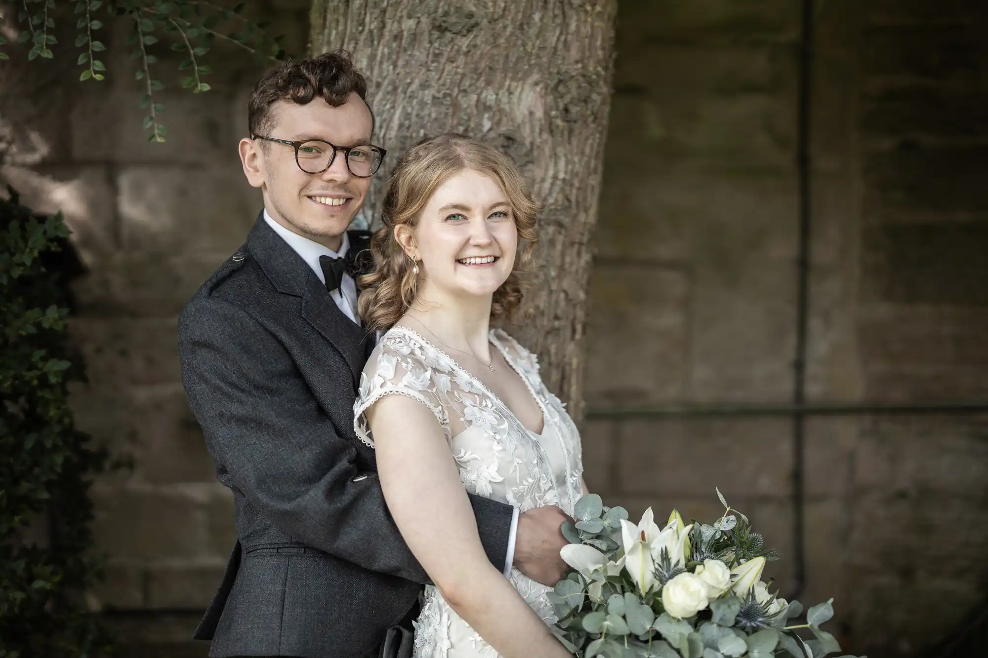 A couple in wedding attire stands by a tree, smiling. The woman holds a bouquet of white flowers and greenery.