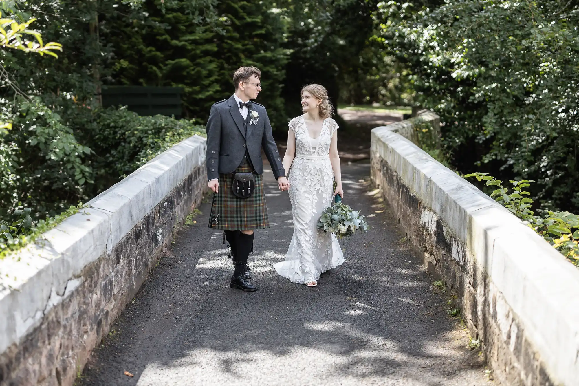 A couple dressed in formal attire, including a kilt for the man and a white dress for the woman, walk hand in hand on a tree-lined stone bridge.