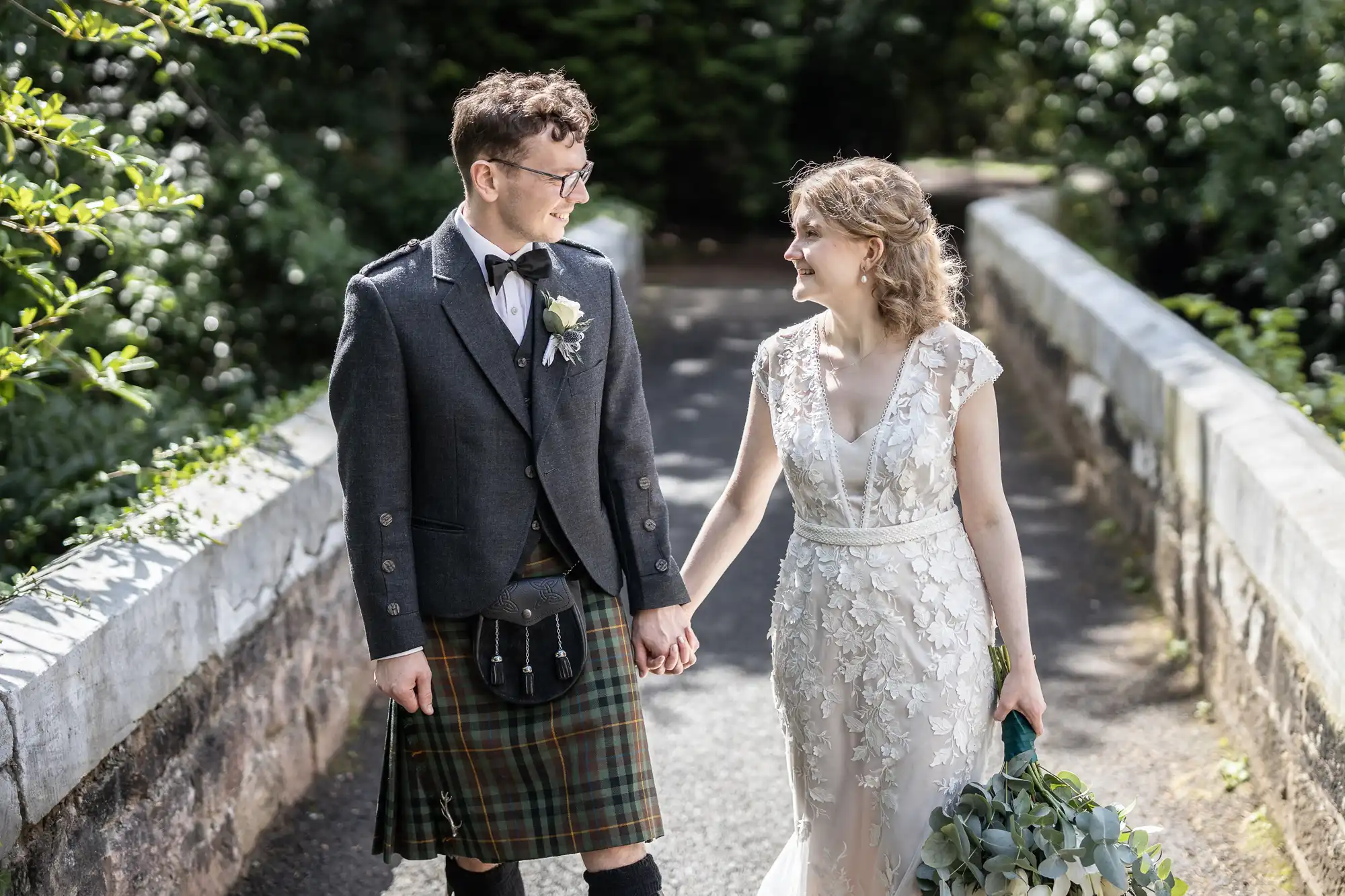 A couple, dressed in wedding attire with the groom in a kilt and the bride in a floral lace gown, walk hand in hand along a stone pathway surrounded by greenery.