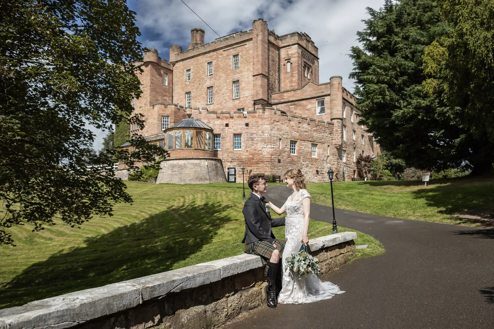 A couple stands beside a stone wall in front of a large historic castle, with the groom in a kilt and the bride in a white dress, holding a bouquet of flowers.