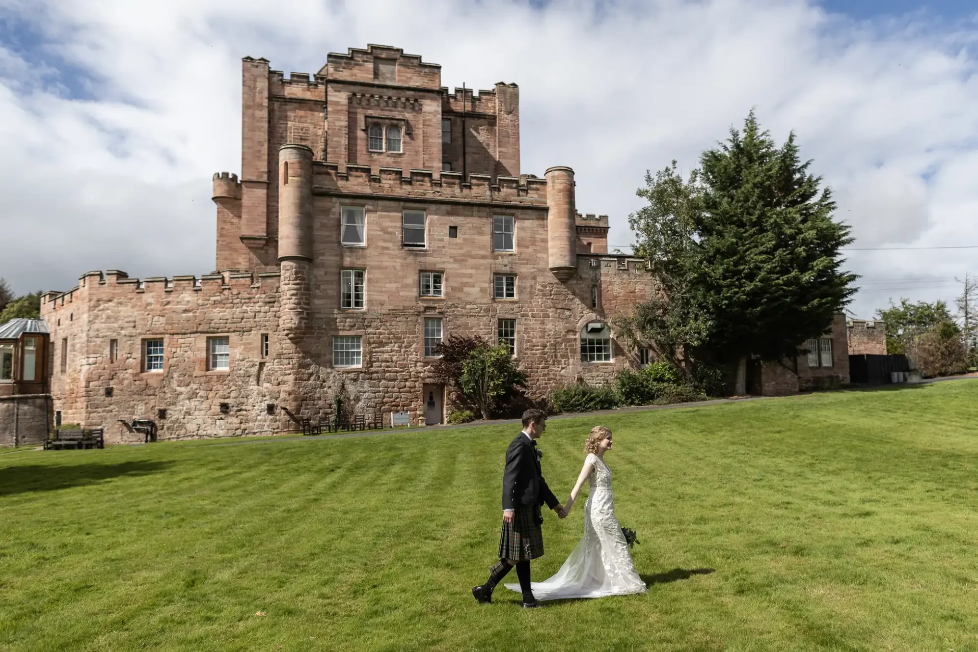 Wedding photography at Dalhousie Castle: a bride and groom walk hand in hand on a lawn in front of a large stone castle under a cloudy sky. The groom is wearing a kilt and the bride is in a white gown.
