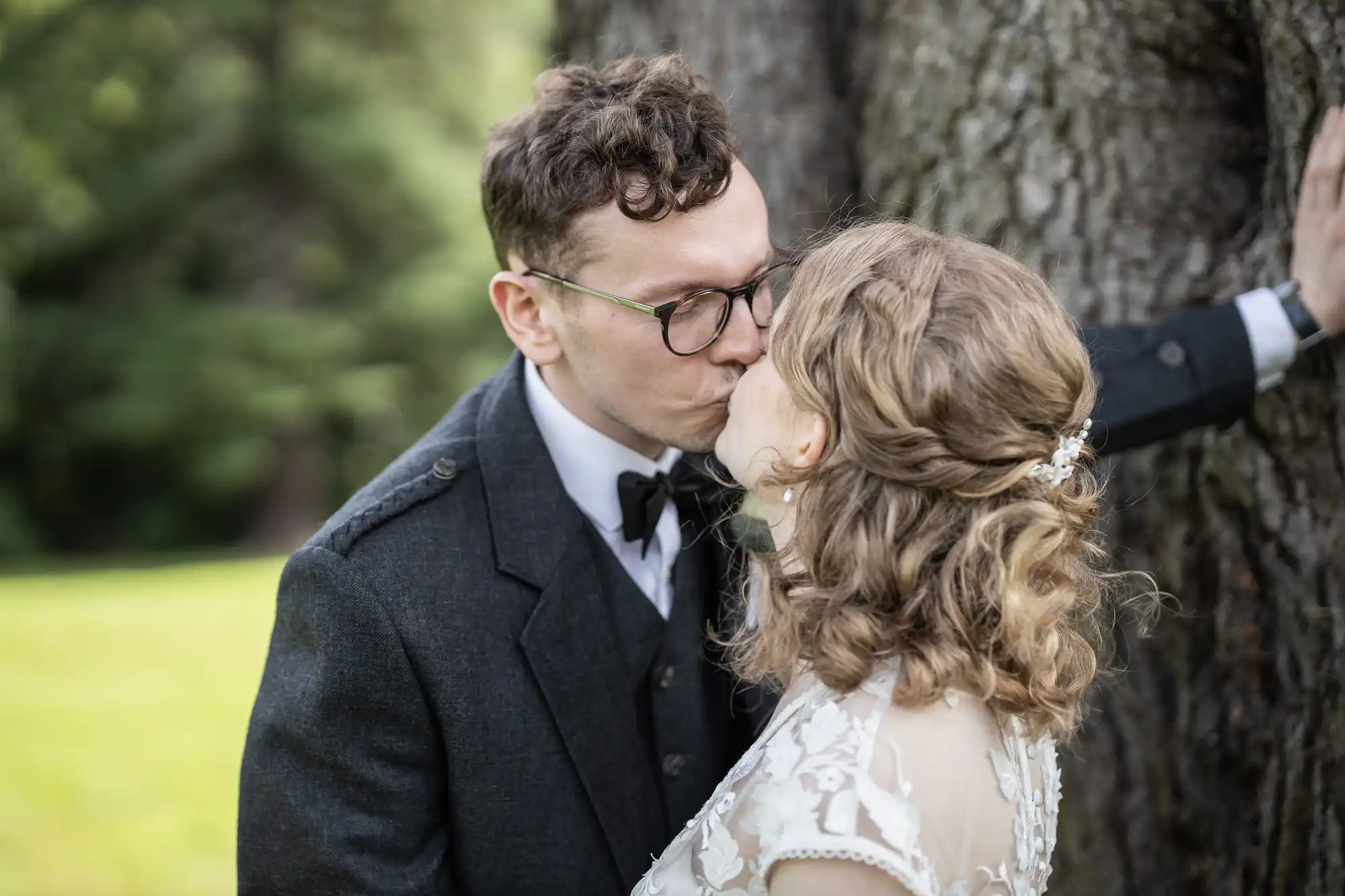 A couple dressed in formal attire share a kiss in front of a large tree outdoors.