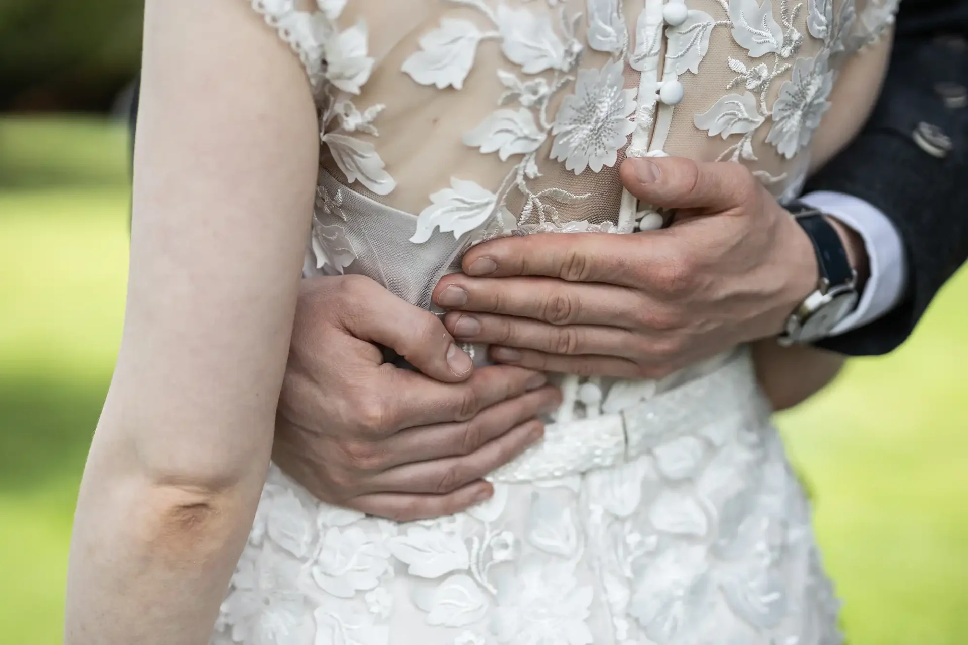 Close-up of a couple embracing; one person wearing a white floral lace dress, the other's hands clasped around their waist, showing a wristwatch.