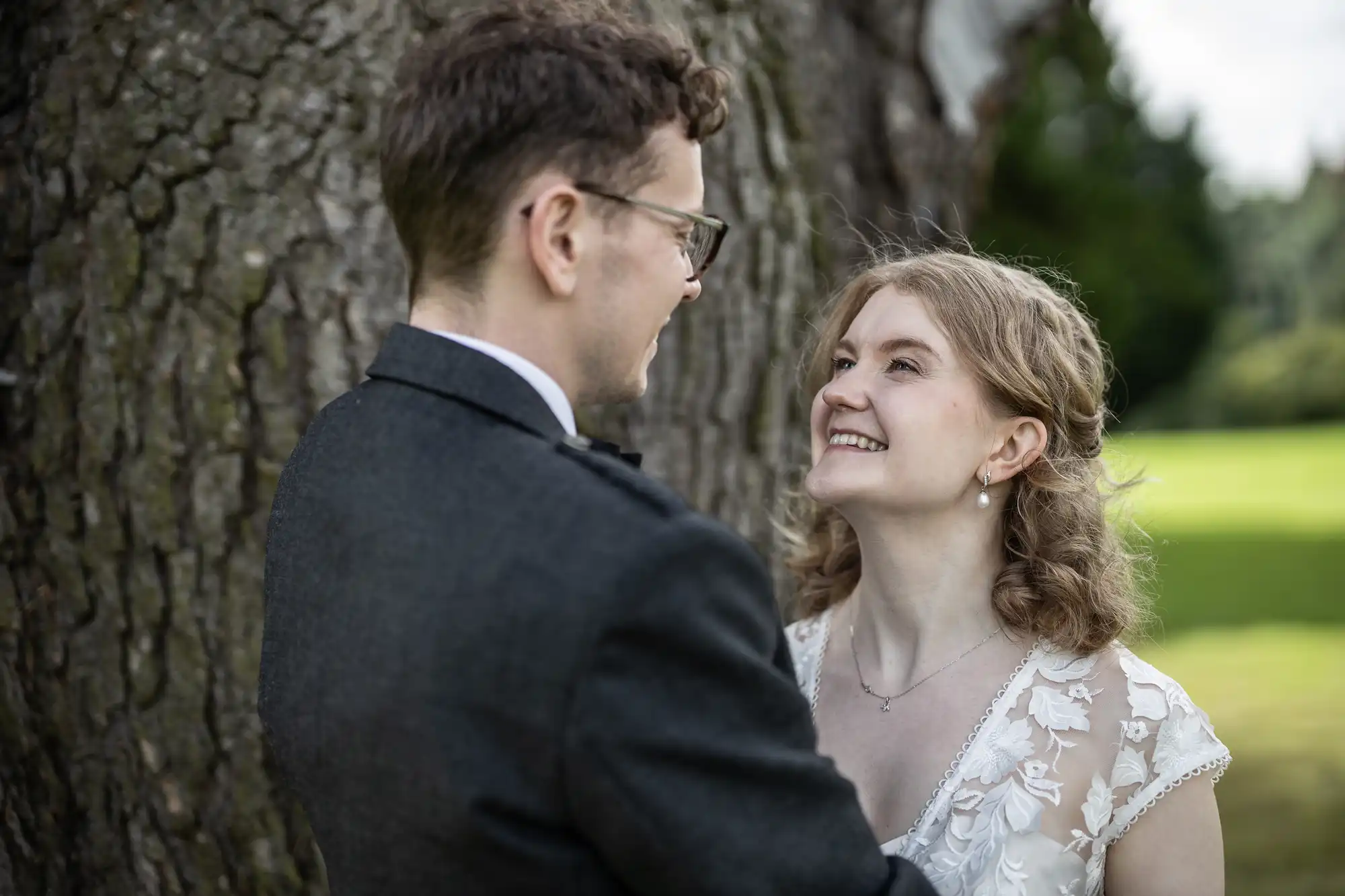 A couple dressed in formal attire smiles at each other while standing outside by a large tree. The background features grass and trees.