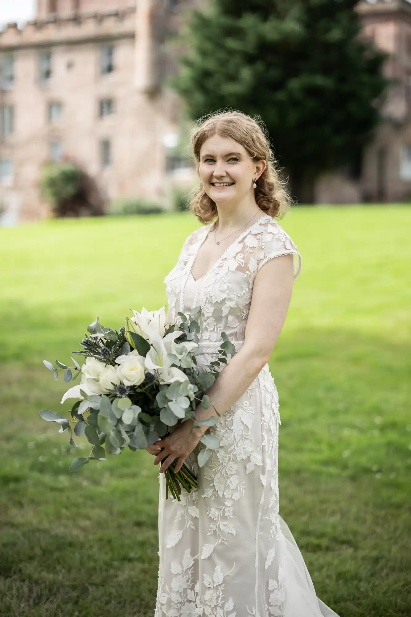 A woman in a white lace gown holding a bouquet of white flowers stands on a grassy area with a building and trees in the background.