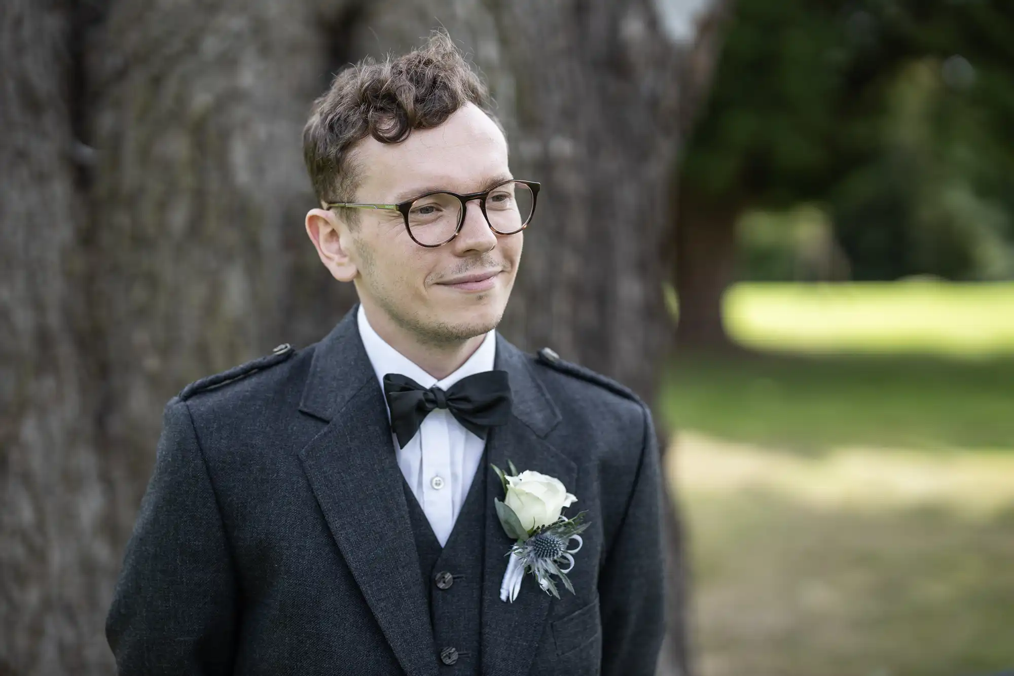 A man with glasses and wavy hair, dressed in a formal dark suit with a bow tie and boutonniere, stands outside near a large tree.