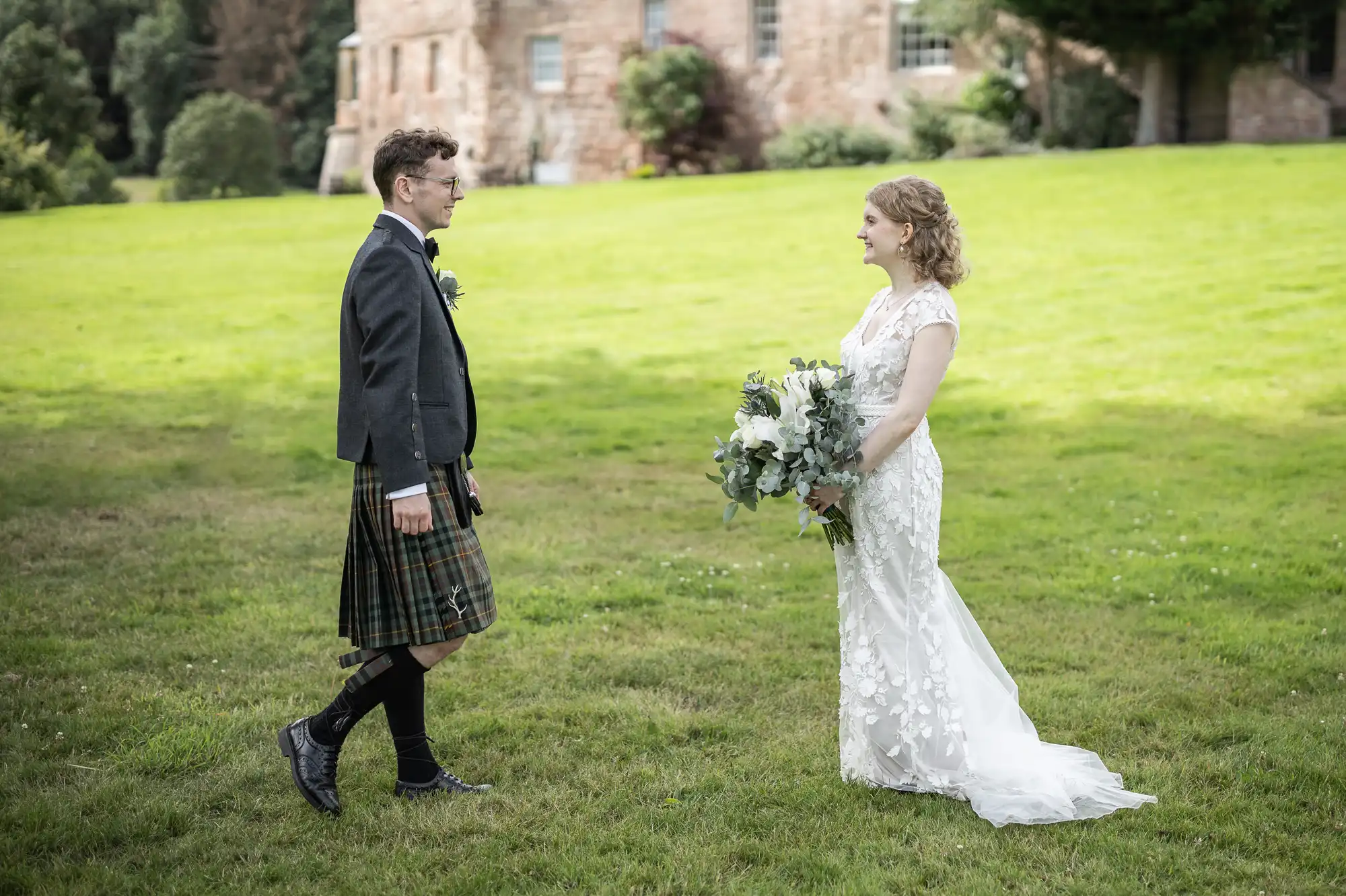 A couple stands facing each other outdoors on a grassy lawn, with an old stone building in the background. The groom is in a kilt; the bride in a white dress holds a large bouquet.