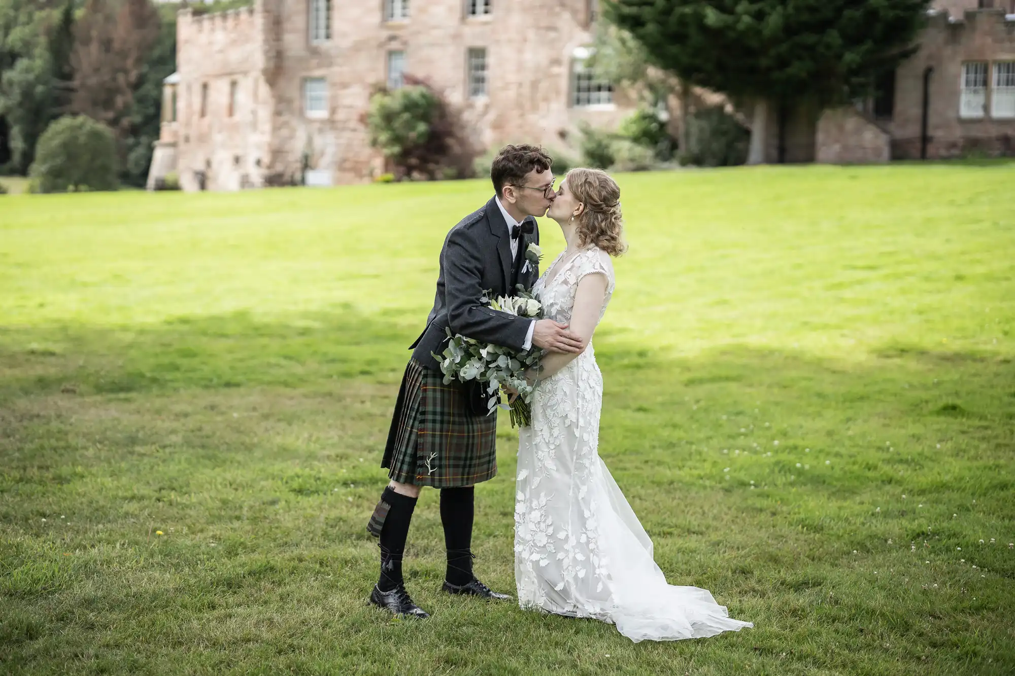 A bride and groom kiss outdoors on a grassy lawn in front of a large stone building. The groom is wearing a kilt, and the bride holds a bouquet.