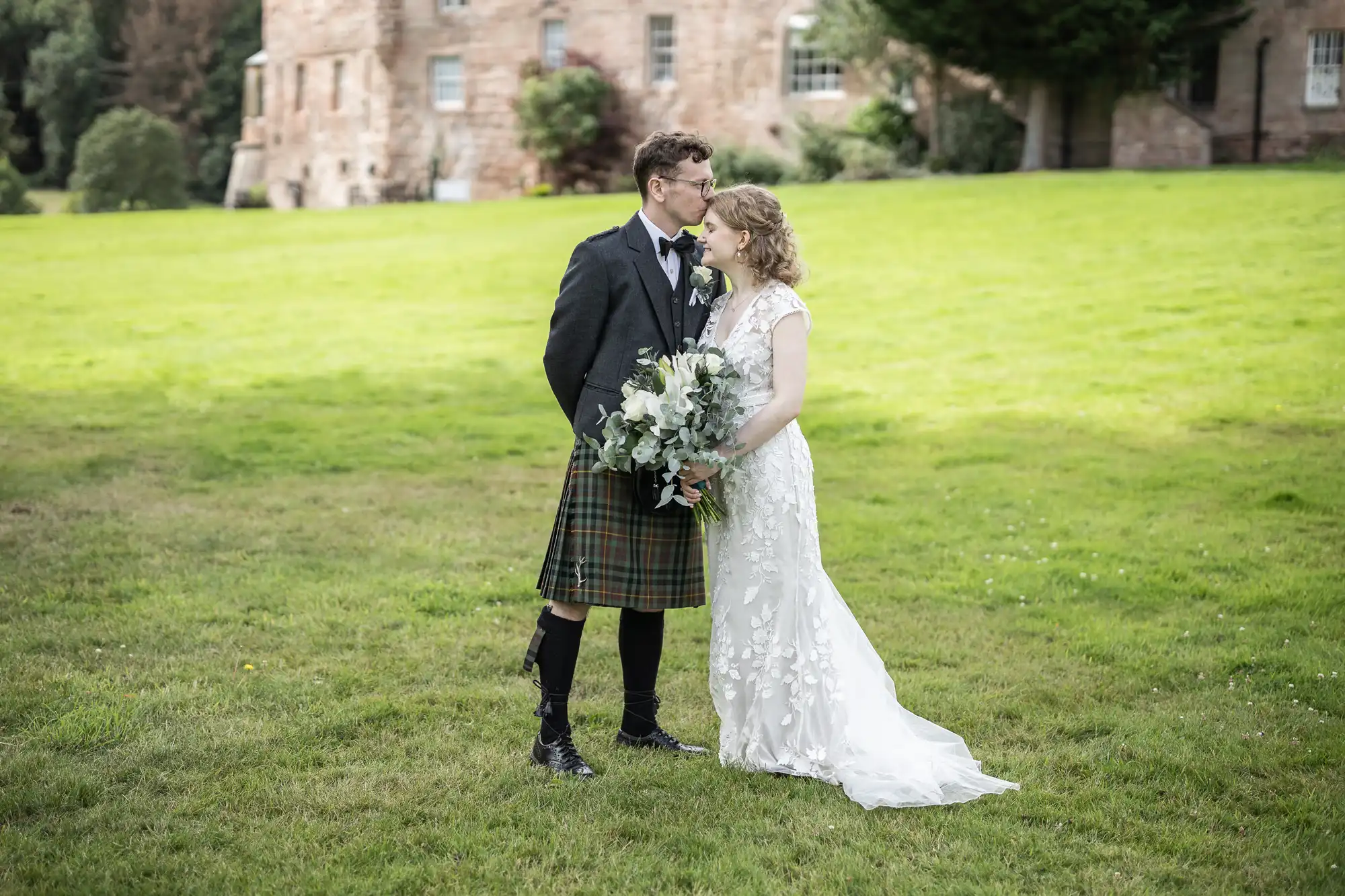 A groom in a kilt and a bride in a white dress stand on a grassy lawn in front of a stone building, embracing and holding a bouquet.