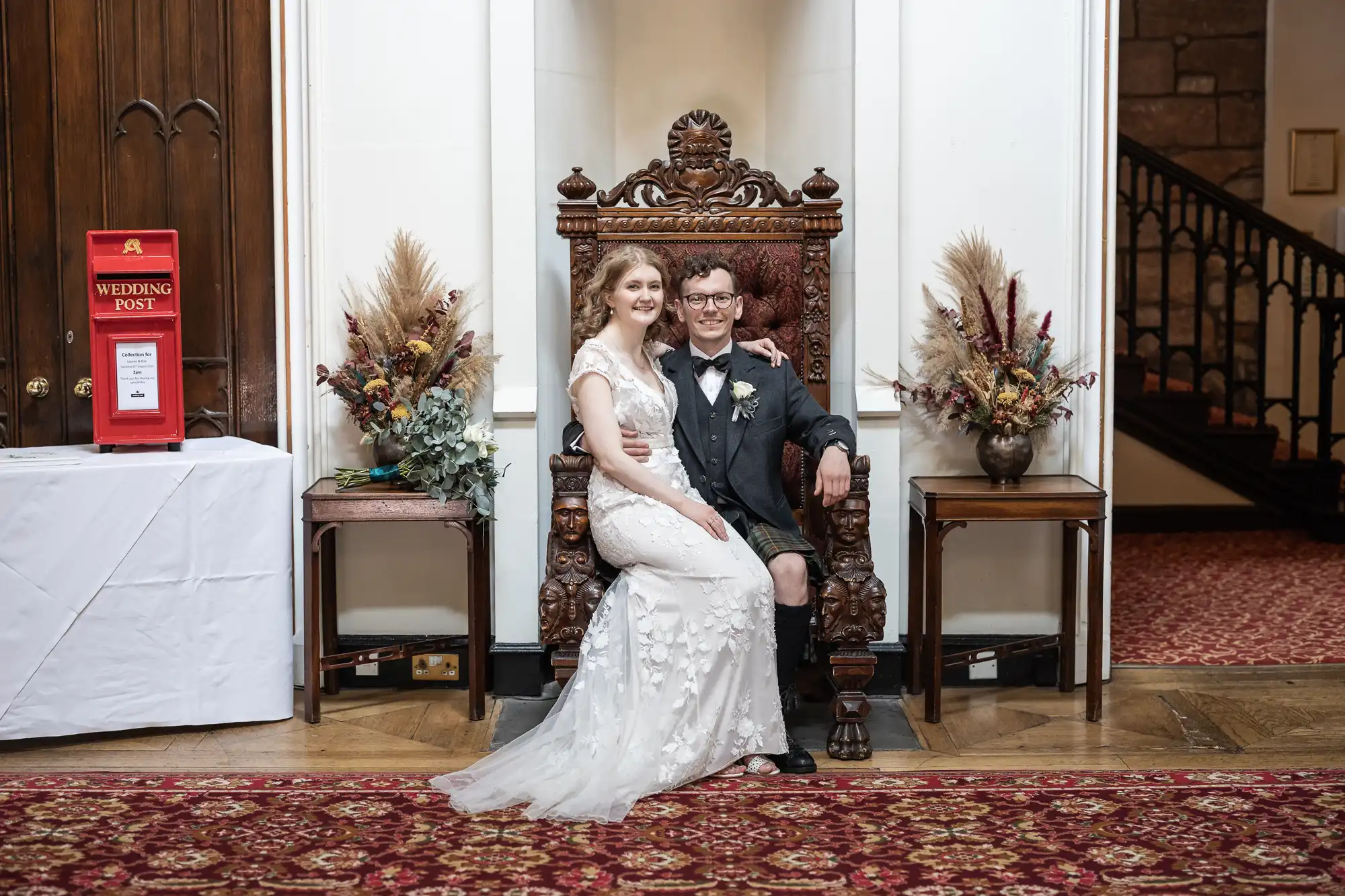 A bride in a white gown sits beside a groom in a suit on a carved wooden chair. They are indoors, flanked by floral arrangements and a red wedding post box.