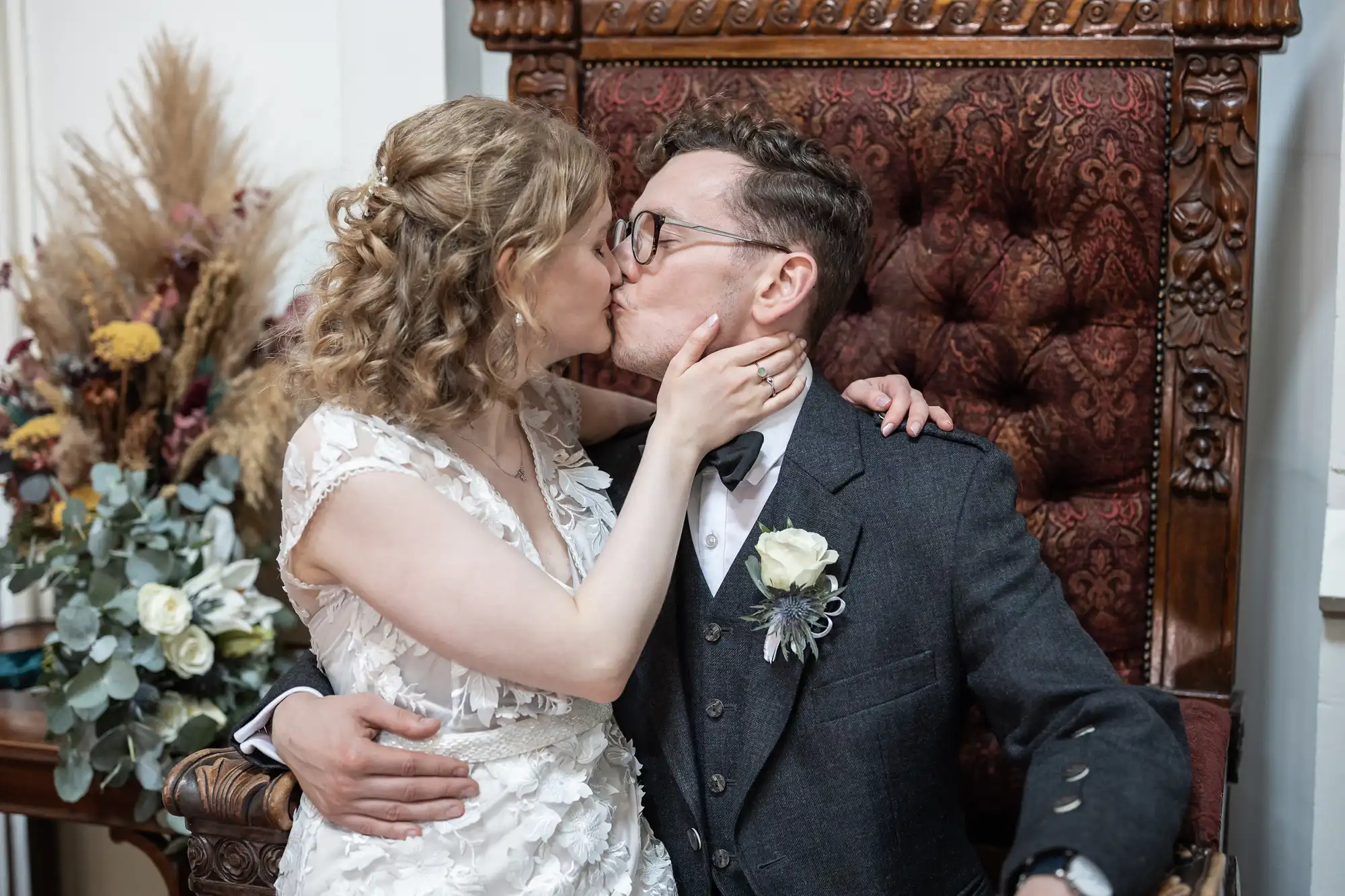 A couple dressed in formal attire kiss while seated on a large, ornate chair. The woman holds the man's face, and there are flowers in the background.