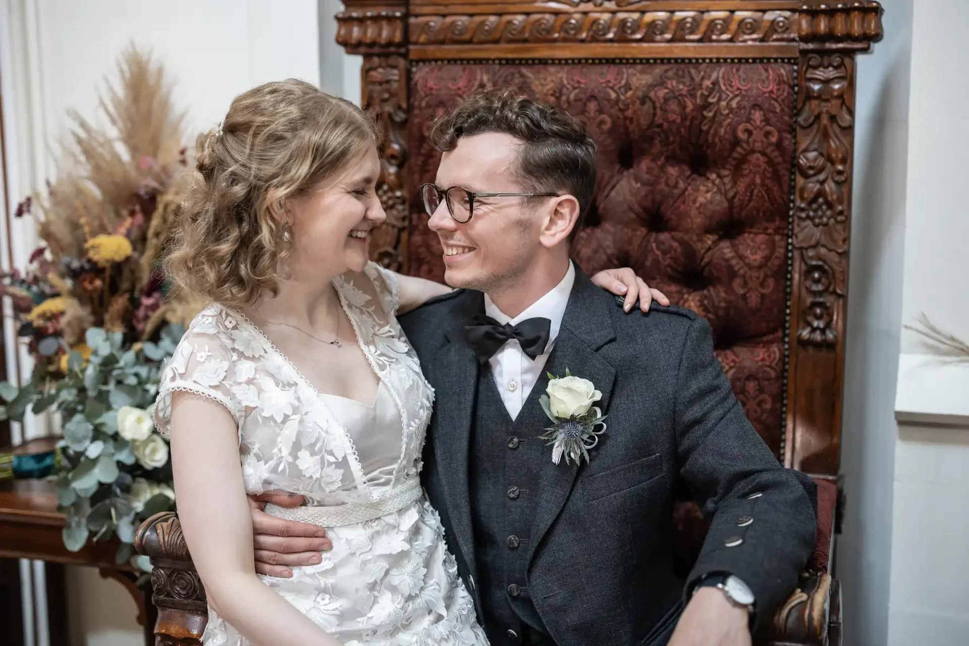 A couple in wedding attire sit on an ornate chair, smiling at each other. The bride wears a white dress, while the groom is dressed in a gray suit with a floral boutonniere. Dried flowers are in the background.