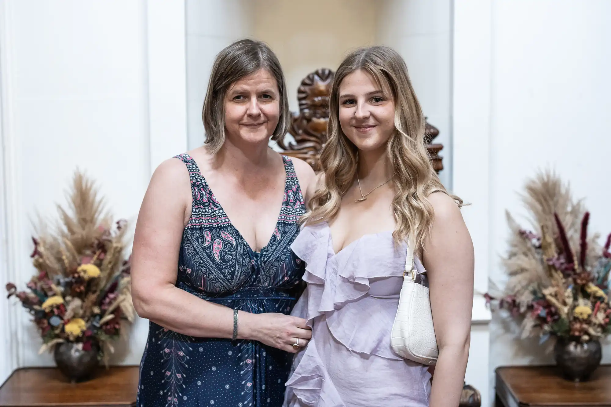 Two women stand together indoors, smiling at the camera. Both are wearing dresses and have their arms around each other. Ornamental floral arrangements are visible in the background.