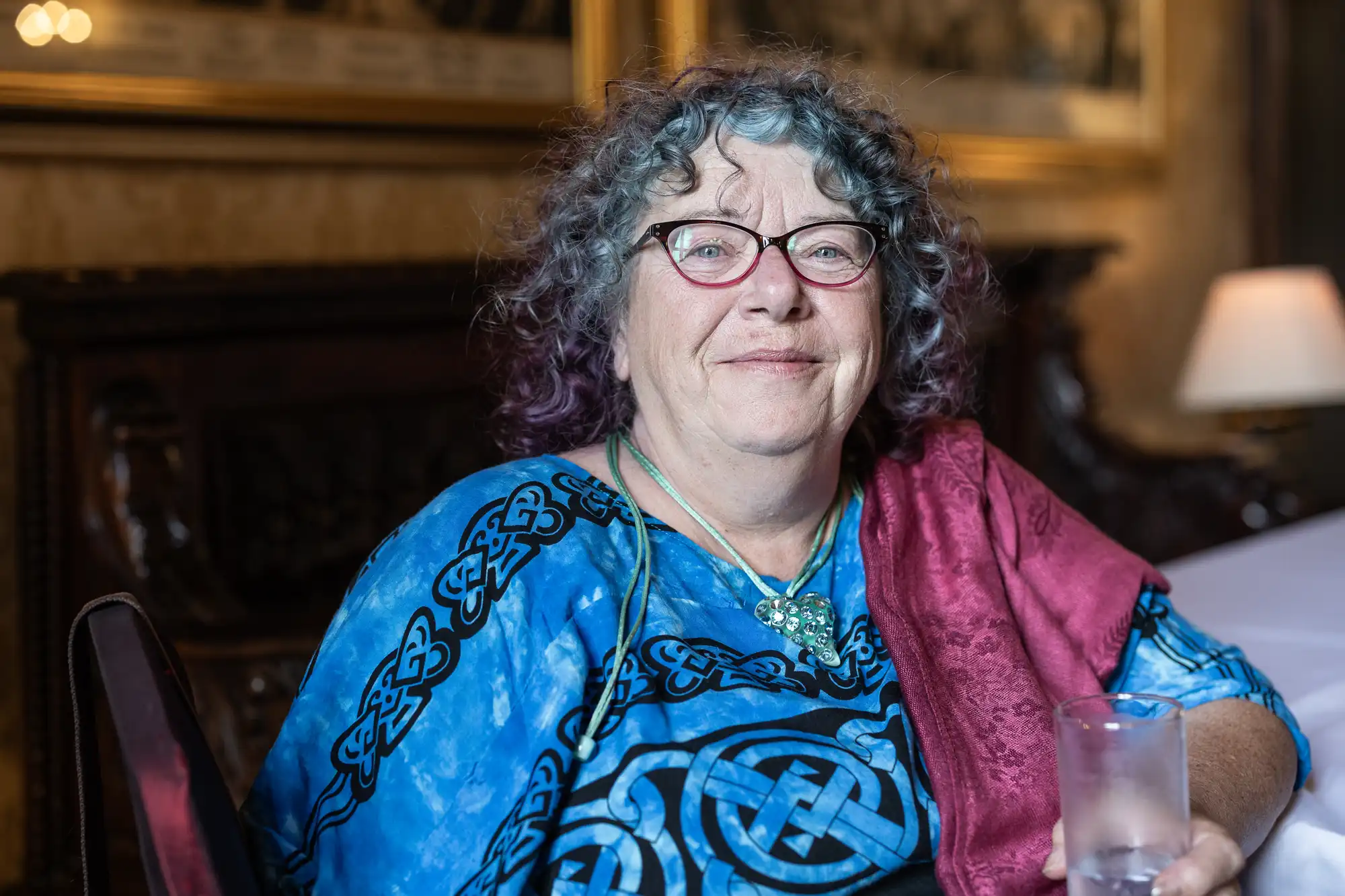 An older woman with curly gray and purple hair, wearing glasses and a blue patterned shirt, sits in a well-decorated room holding a glass of water.