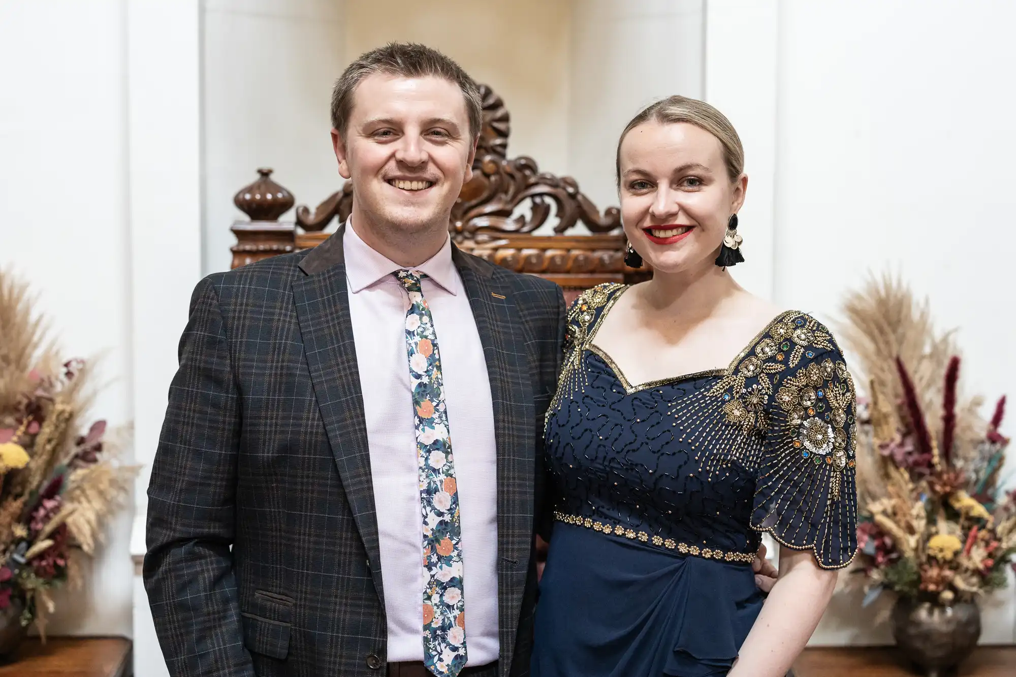 A man in a checked suit and floral tie stands next to a woman in an ornate navy dress, posing for a photo indoors with floral arrangements in the background.