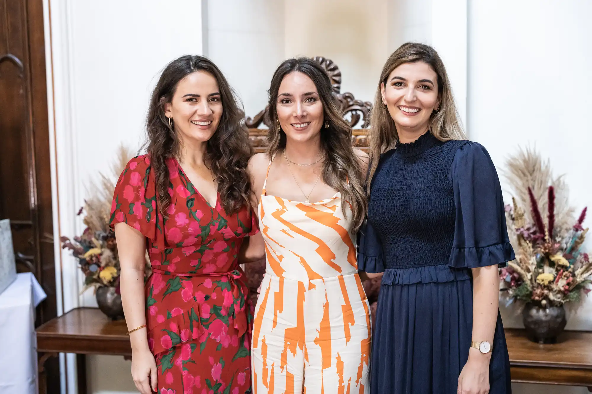 Three women stand close together smiling, dressed in vibrant attire, with a decorative background featuring floral arrangements.